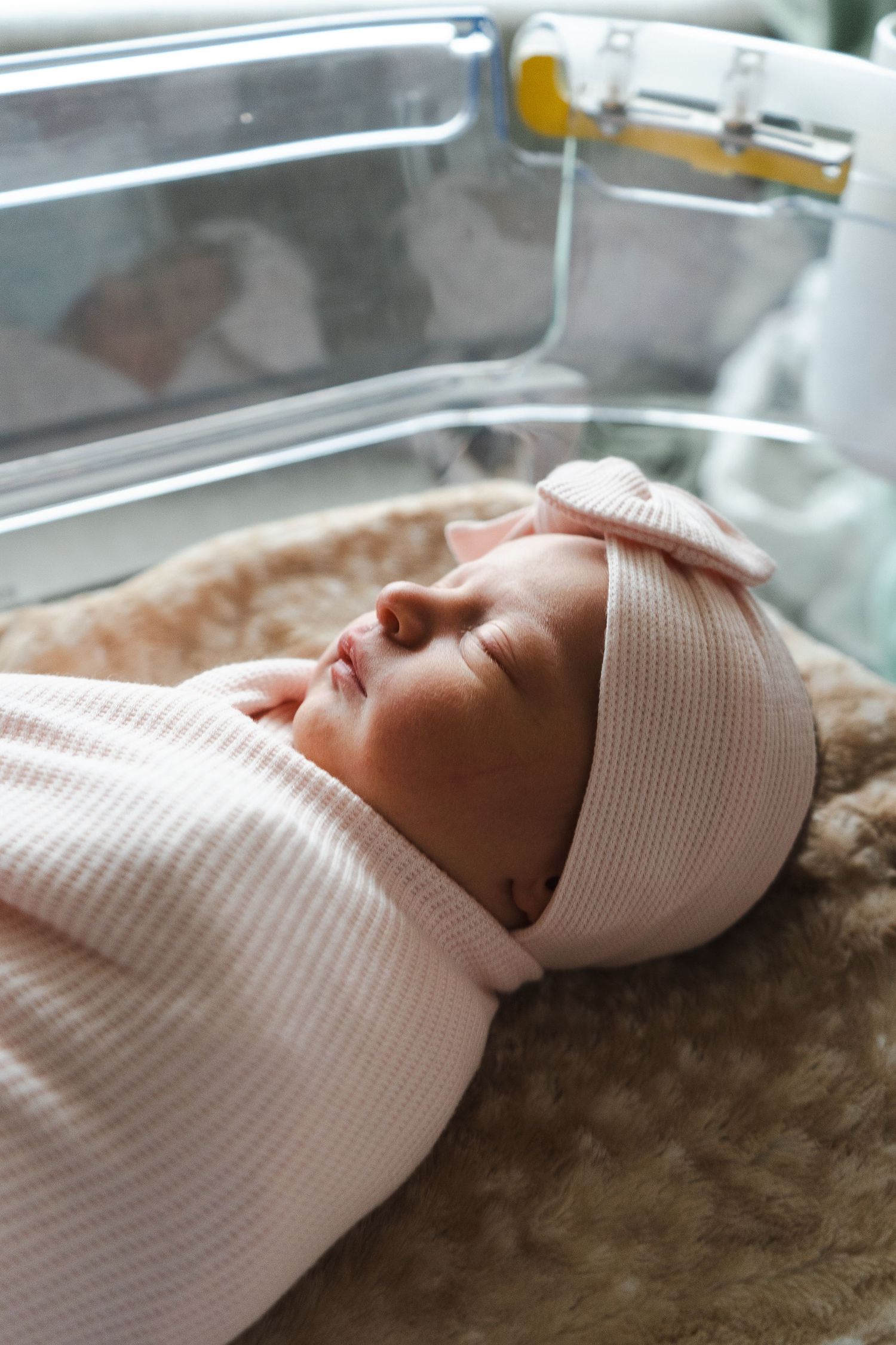 A baby rests peacefully in a hospital bassinet wearing a pink and white striped blanket and white cap.