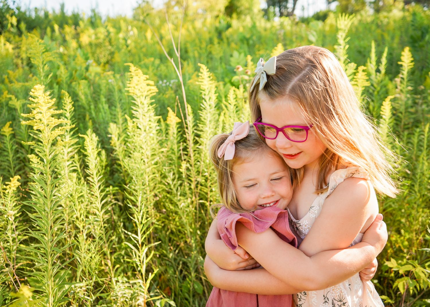 Sisters sharing a warm embrace while standing in a sunny meadow with golden vegetation.