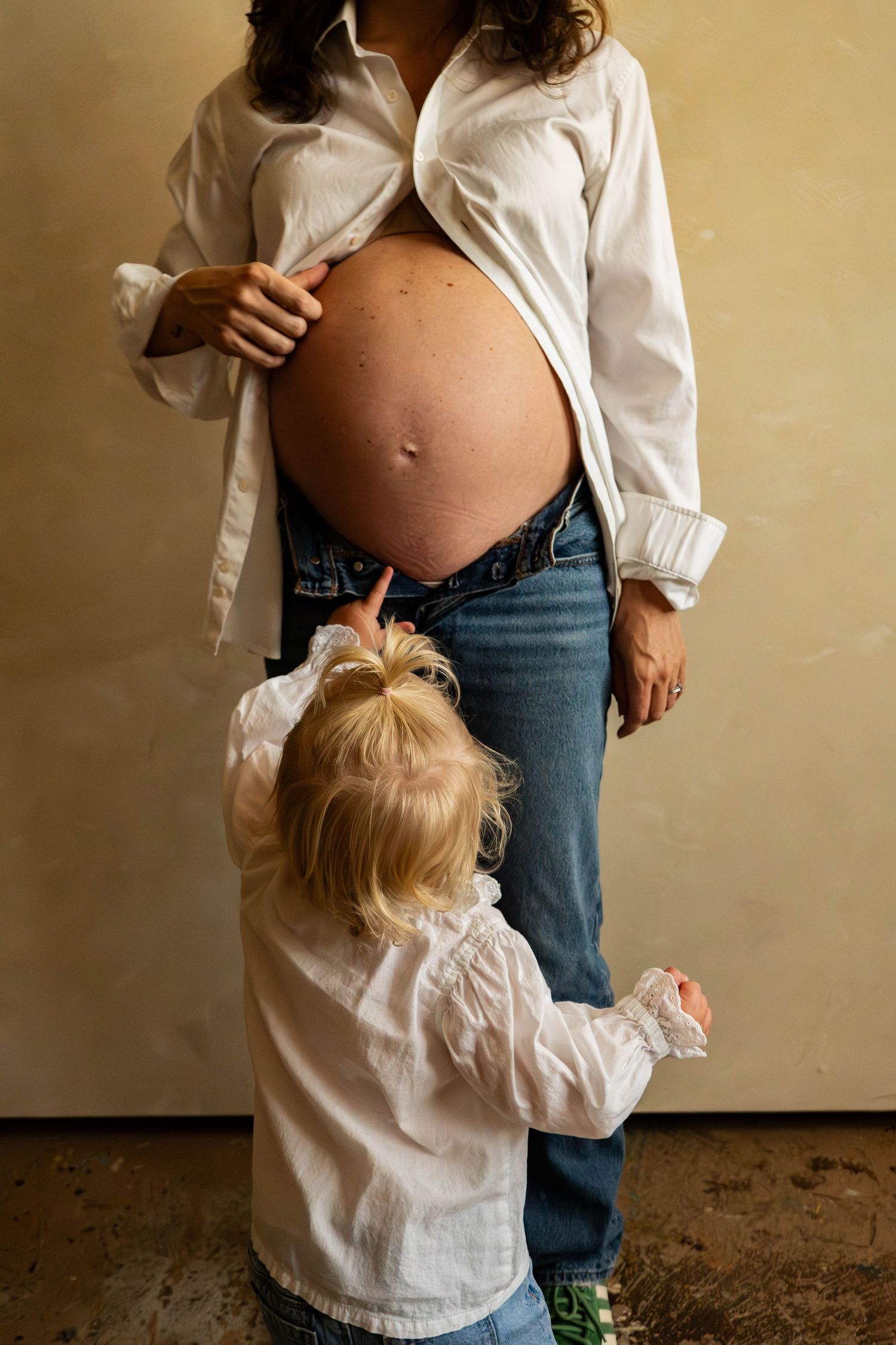 Sweet maternity moment with toddler reaching toward pregnant belly in matching white shirts.