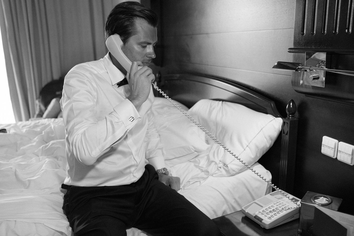 A black and white photo of someone in formal attire making a phone call in a hotel room.