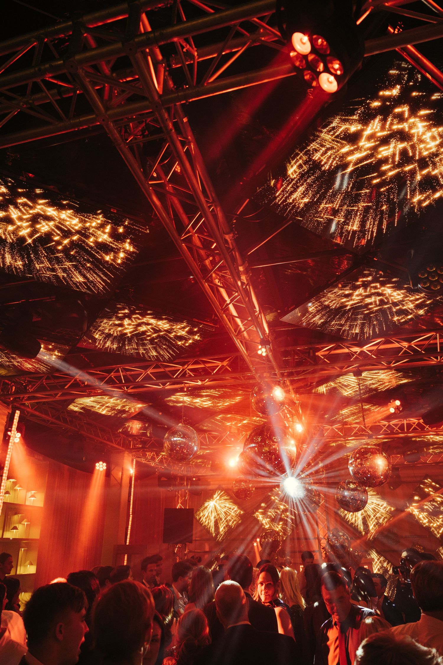 Dramatic red stage lighting and golden sparklers illuminate a crowded nightclub dance floor.