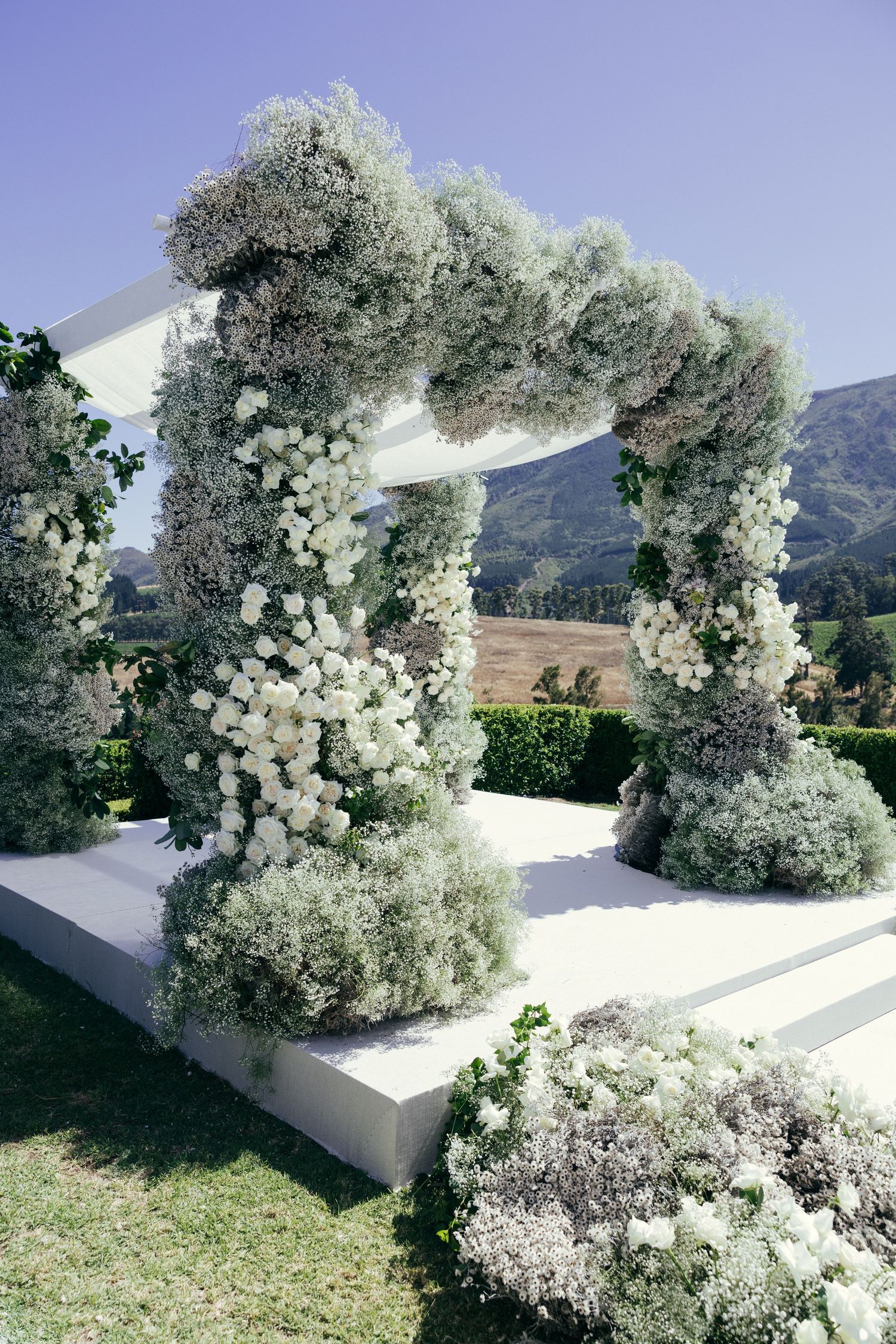 White floral wedding arch with baby's breath and roses against mountain backdrop.