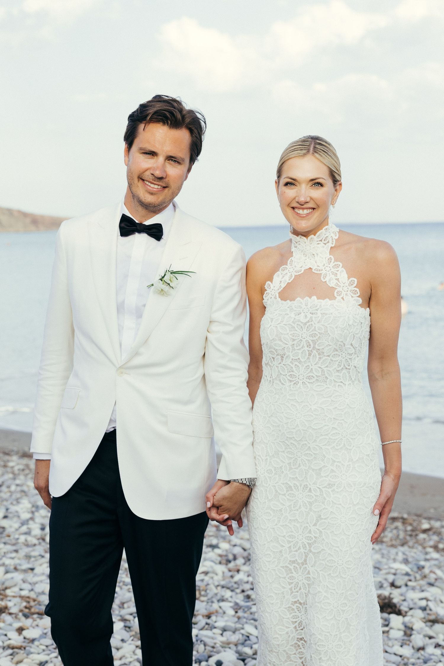 Wedding couple poses together on a rocky beach wearing white formal attire with ocean waves in background.