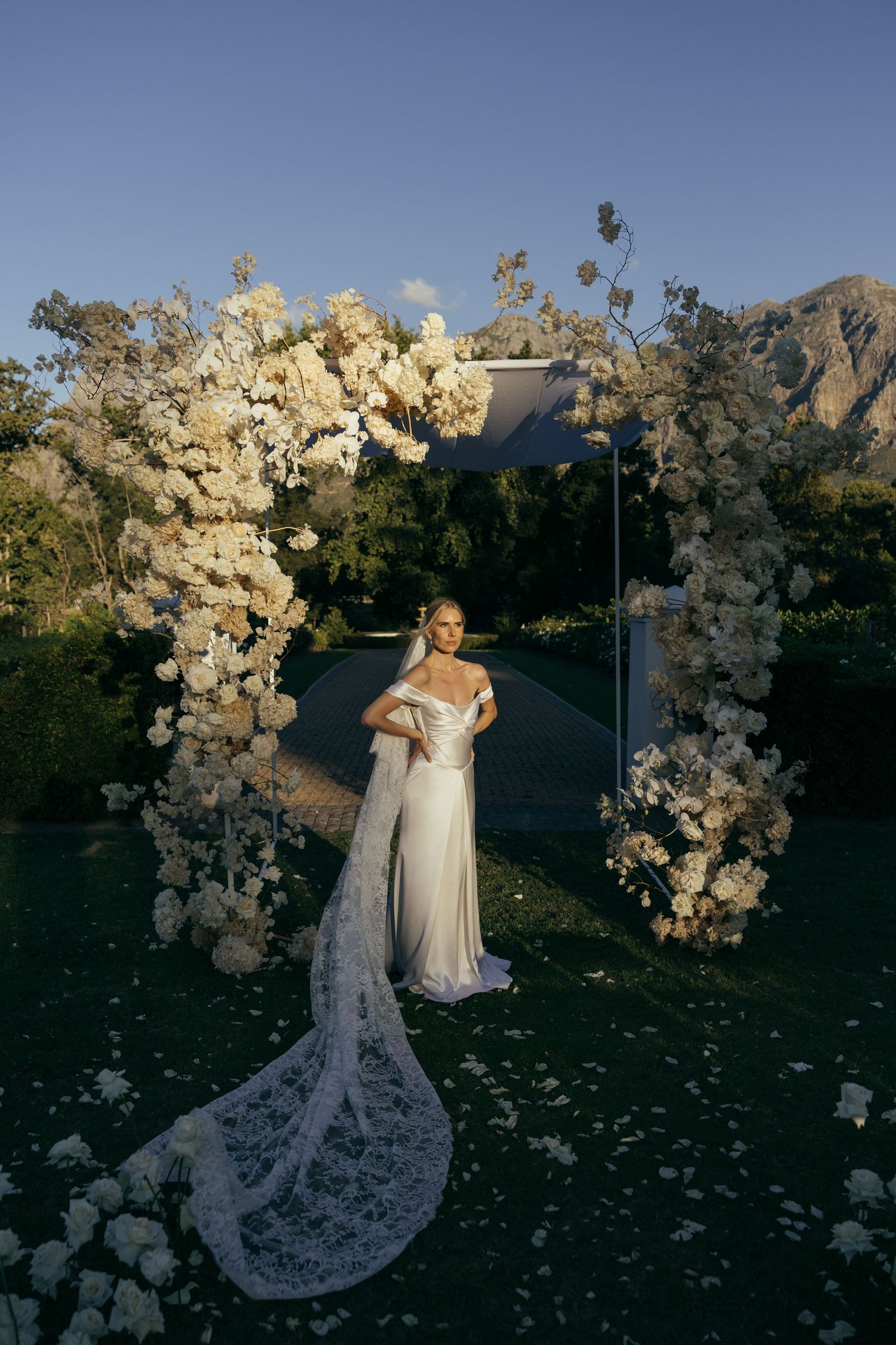 Wedding couple poses in front of white floral arch with mountain landscape backdrop.
