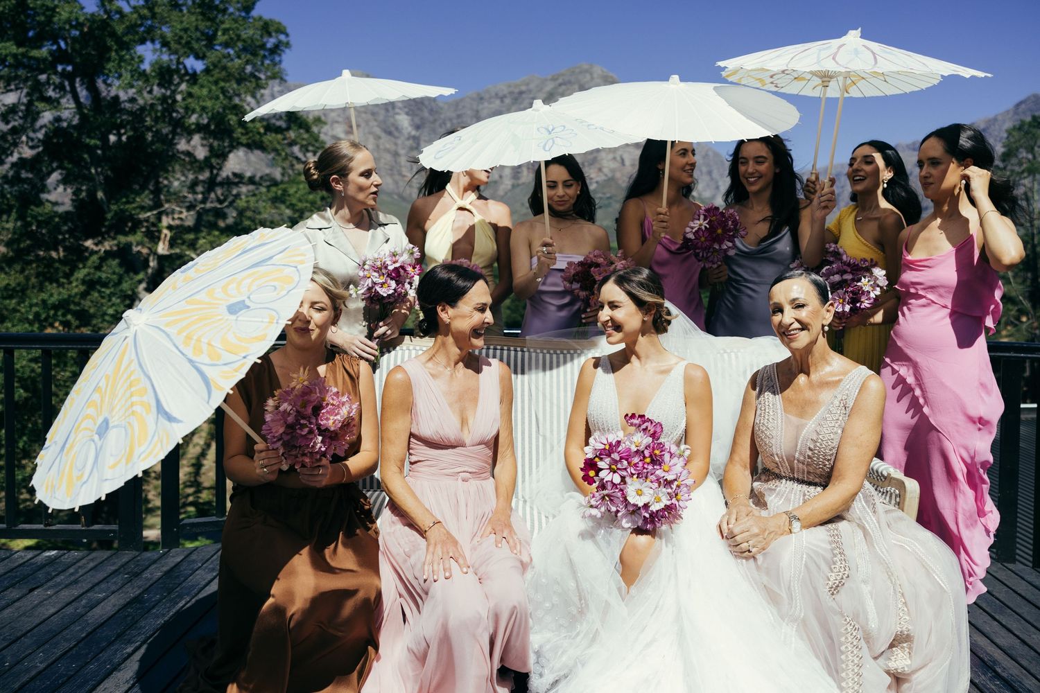 Wedding party with parasols gathered on a sunny deck wearing pink dresses.