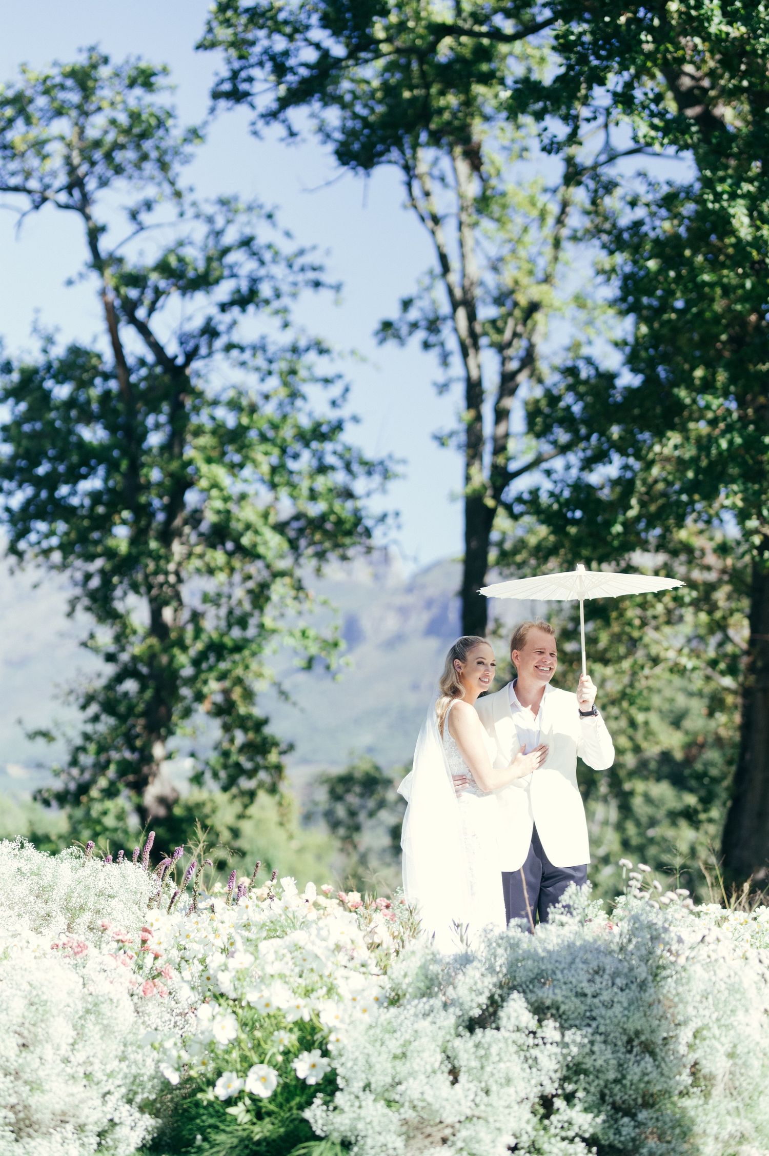 A couple stands under a white umbrella surrounded by delicate white flowers in a garden setting.