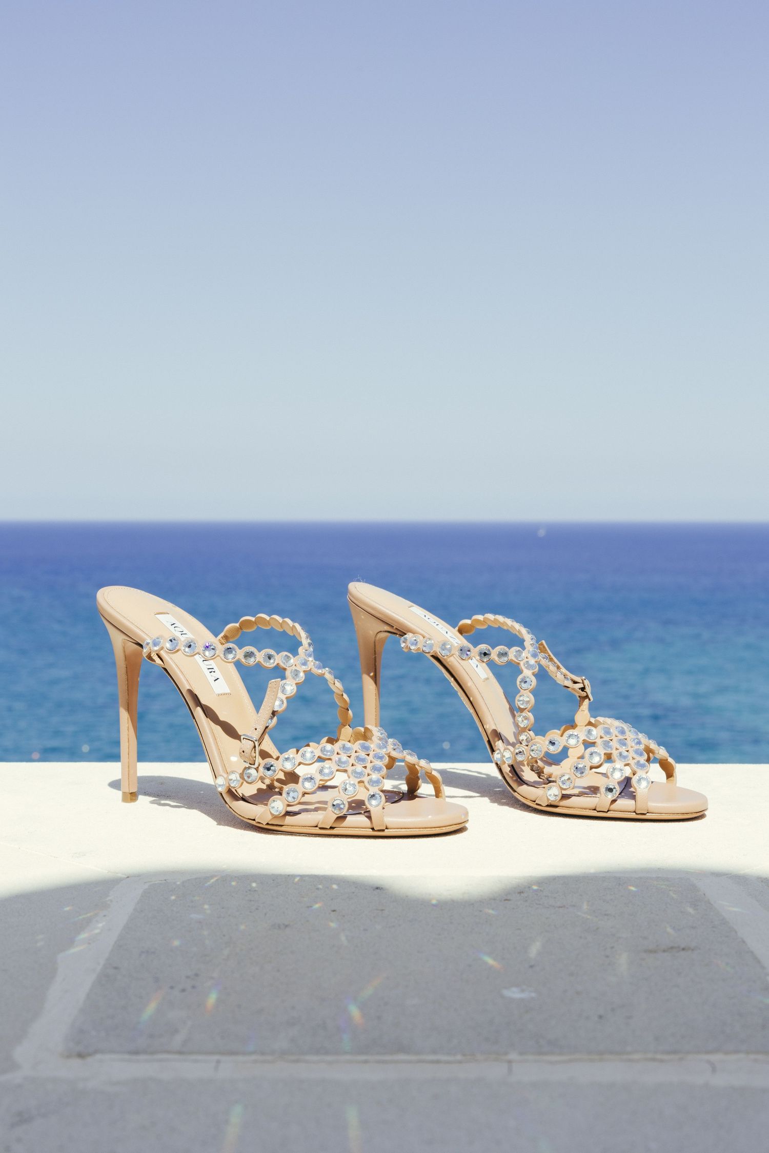 Elegant jeweled bridal sandals displayed against an ocean backdrop on a sunny day.