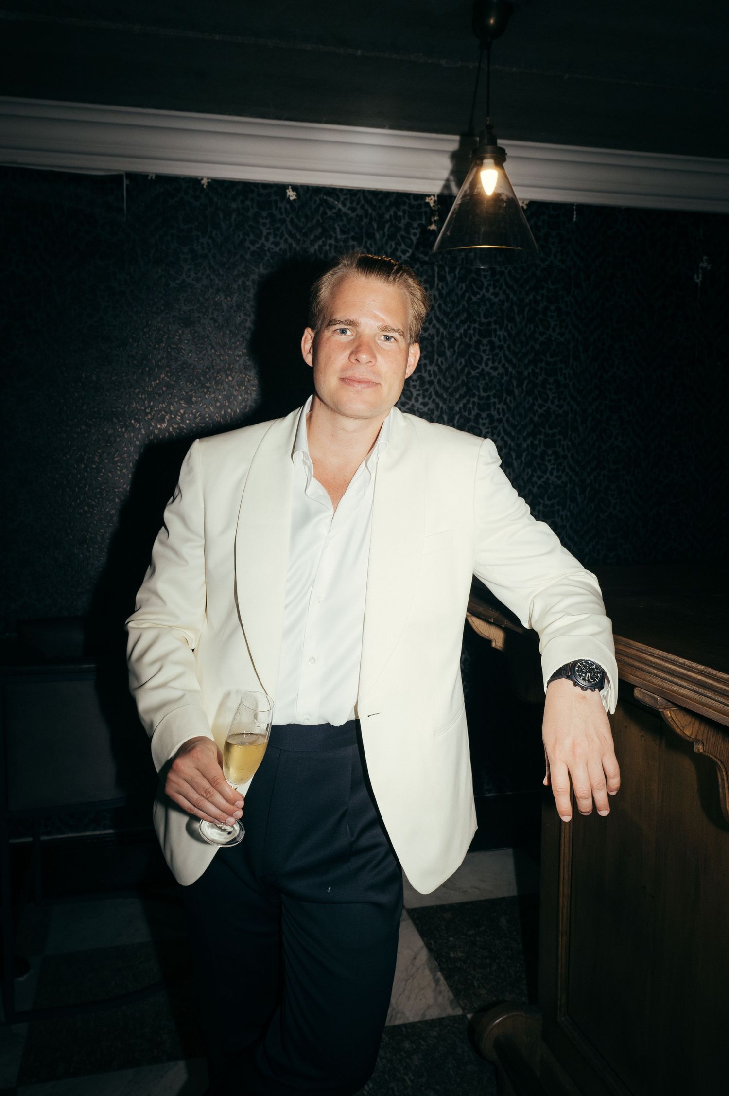 A person in a white blazer and dark pants holds a drink in a dimly lit room.