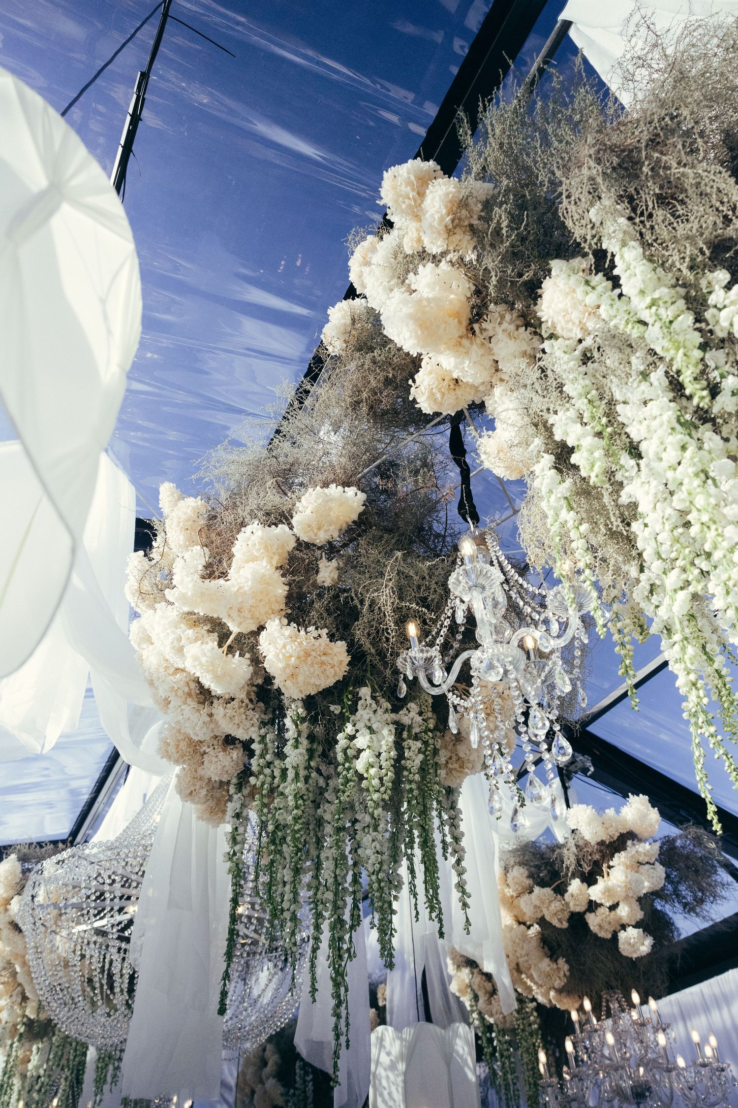 Luxurious wedding decor featuring white floral arrangements and crystal chandeliers under a clear tent.