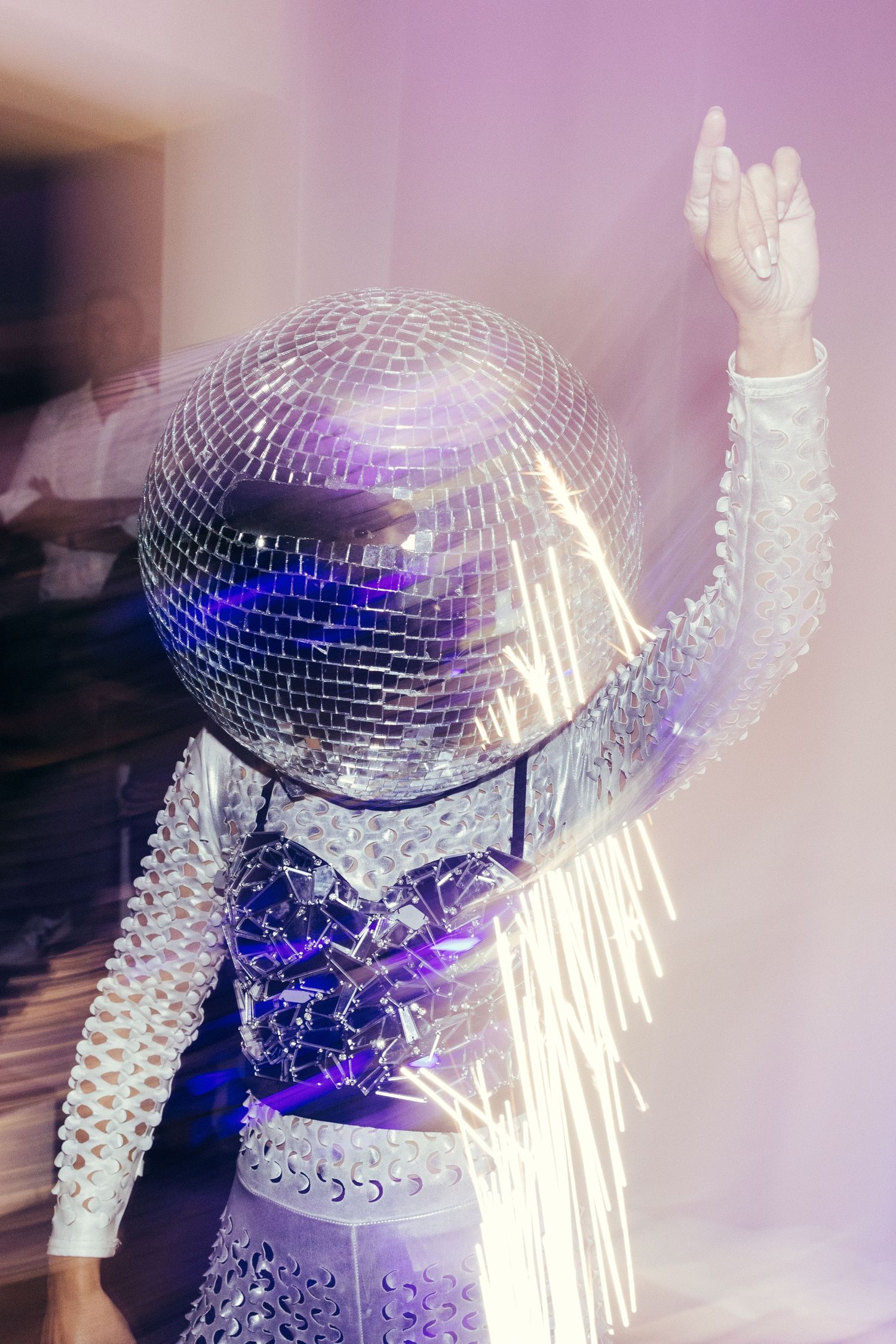 A figure in white sequins poses with a disco ball head in purple lighting.