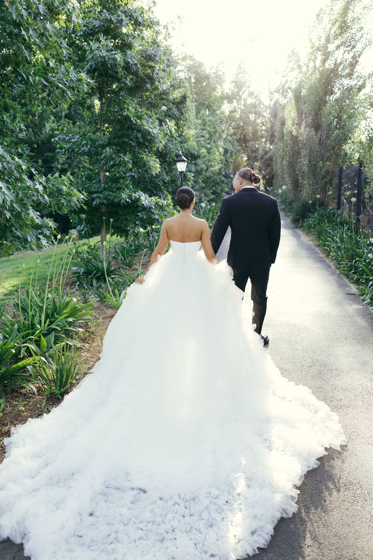 Newlyweds walking hand in hand down a sunlit garden path with a dramatic flowing wedding dress train.
