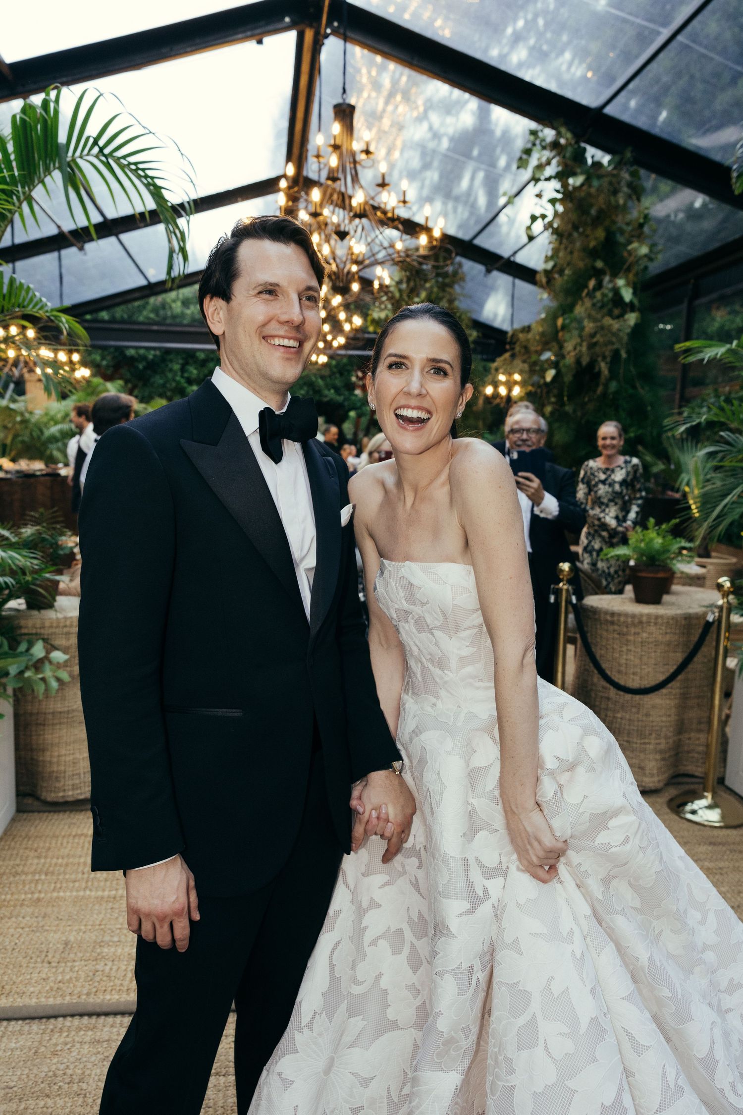 Wedding couple shares joyful moment in greenhouse venue decorated with string lights and tropical plants.