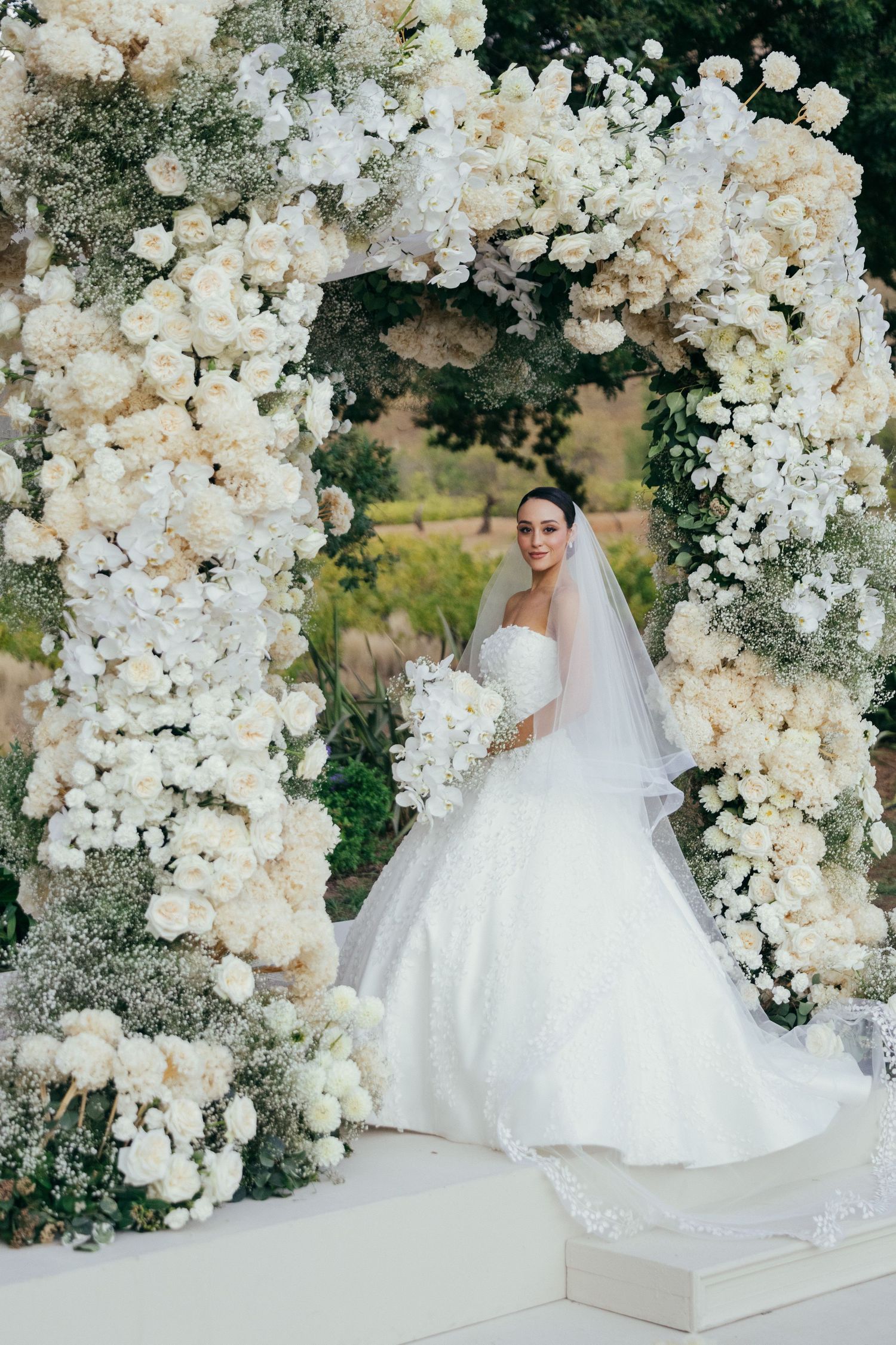 Luxurious white floral wedding arch with cascading roses and orchids forms a romantic ceremony backdrop.