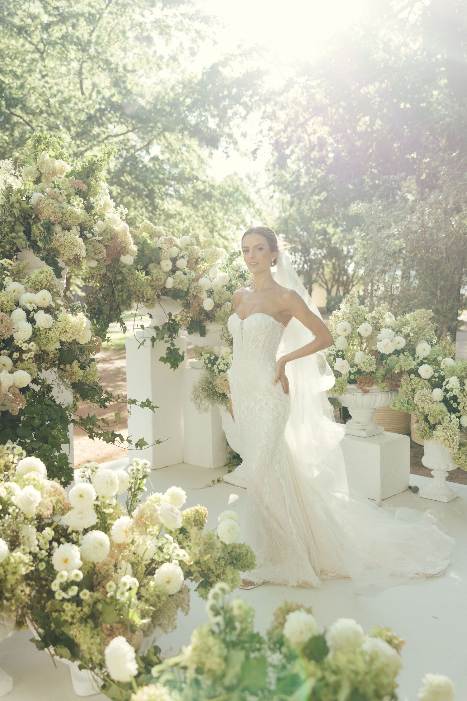 Dreamy garden wedding setup with white floral arrangements and columns in natural sunlight.