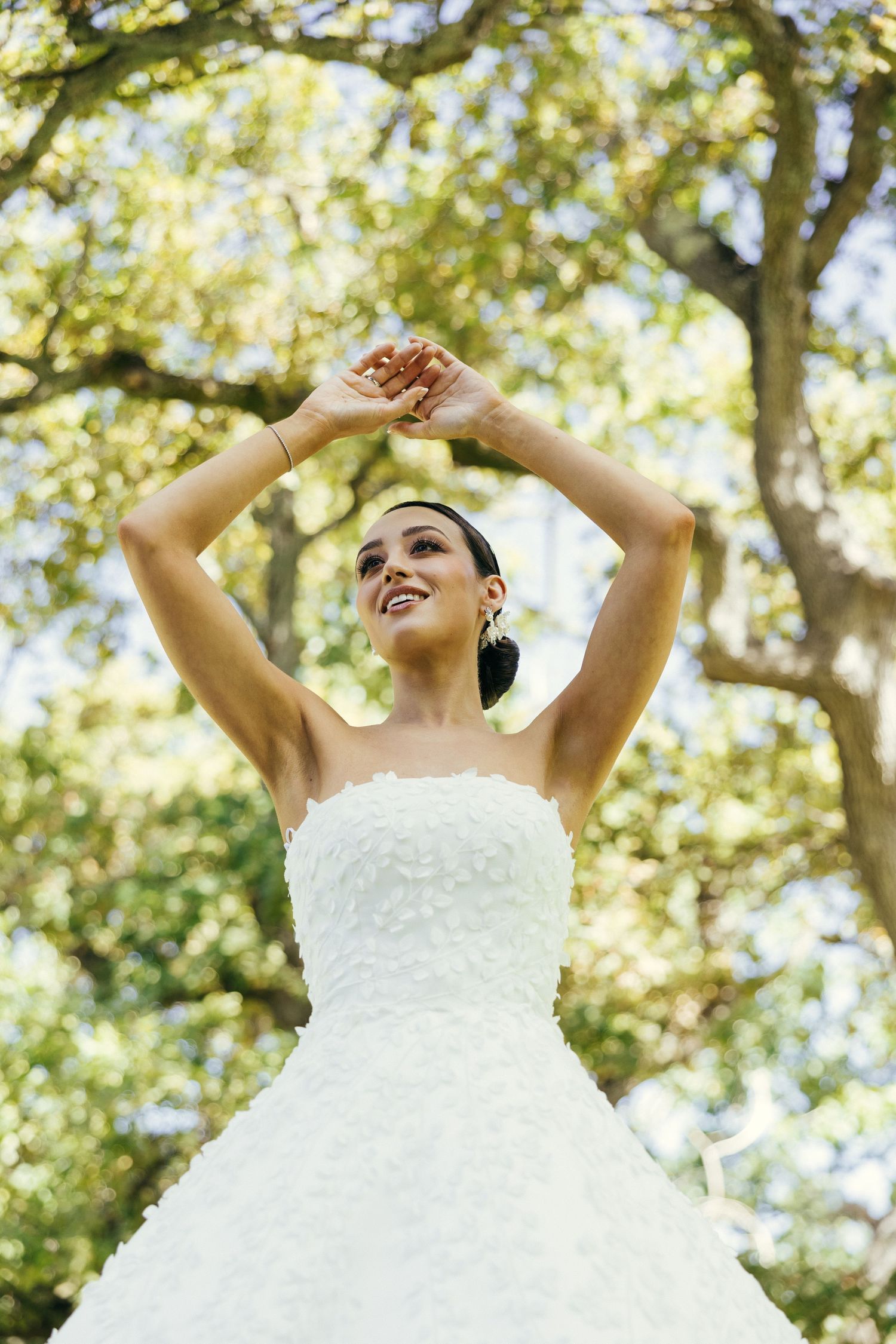Joyful bride in strapless gown raises arms under sunlit trees.
