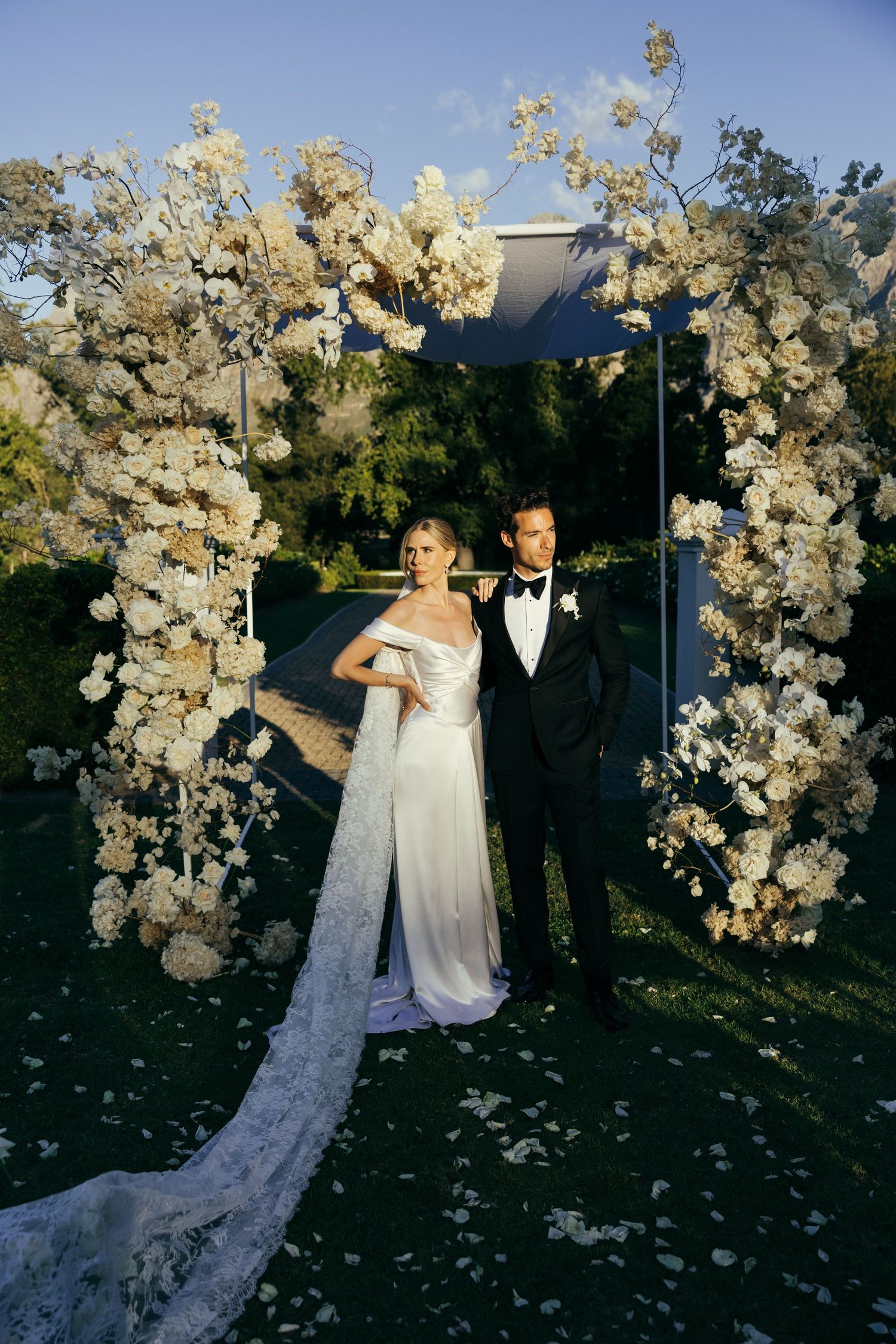 Romantic wedding photoshoot with dramatic white floral arch against blue sky and mountains.