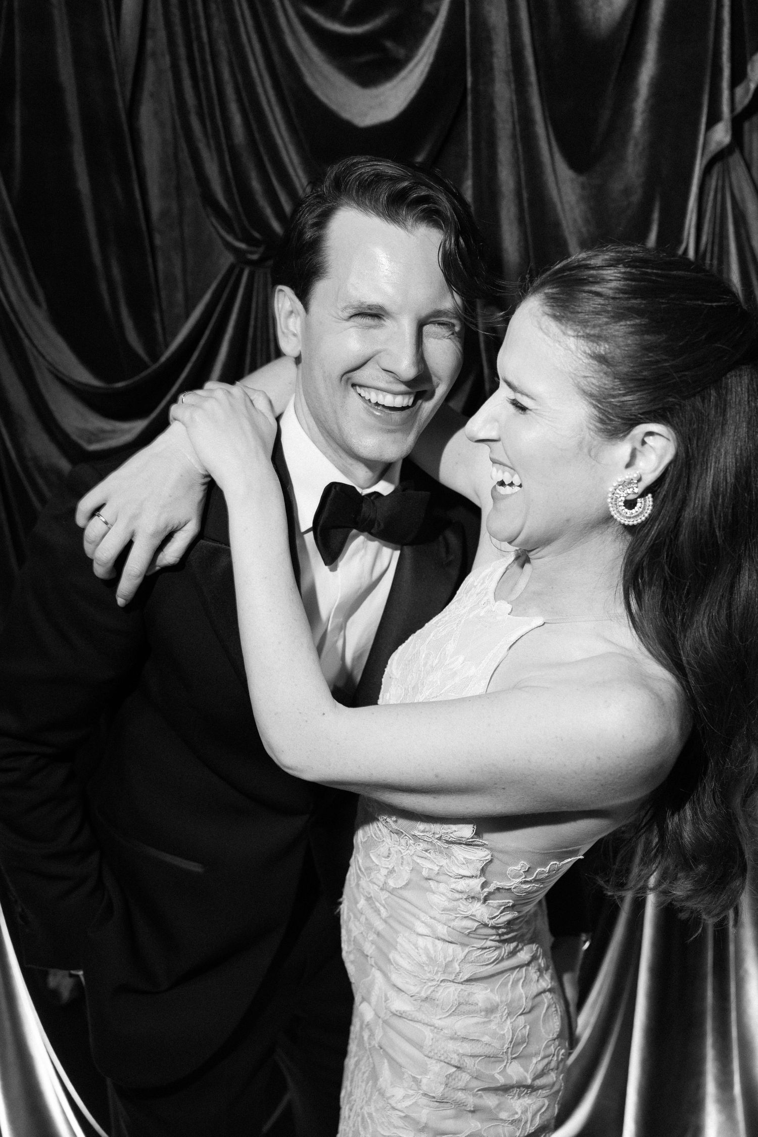 Black and white portrait of newlyweds laughing together against dramatic fabric backdrop.