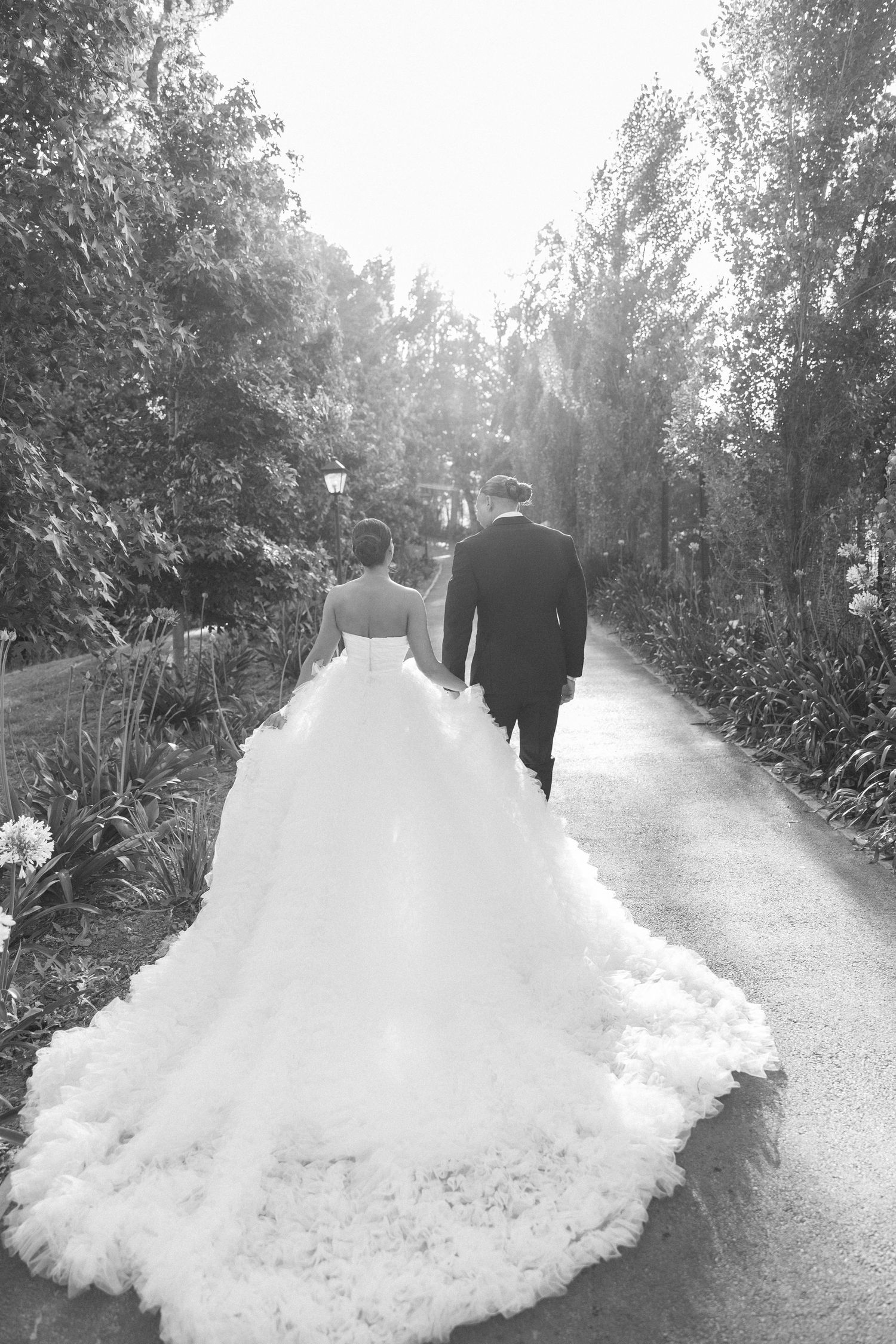 Black and white photo of a wedding couple walking down a garden path with a flowing train.