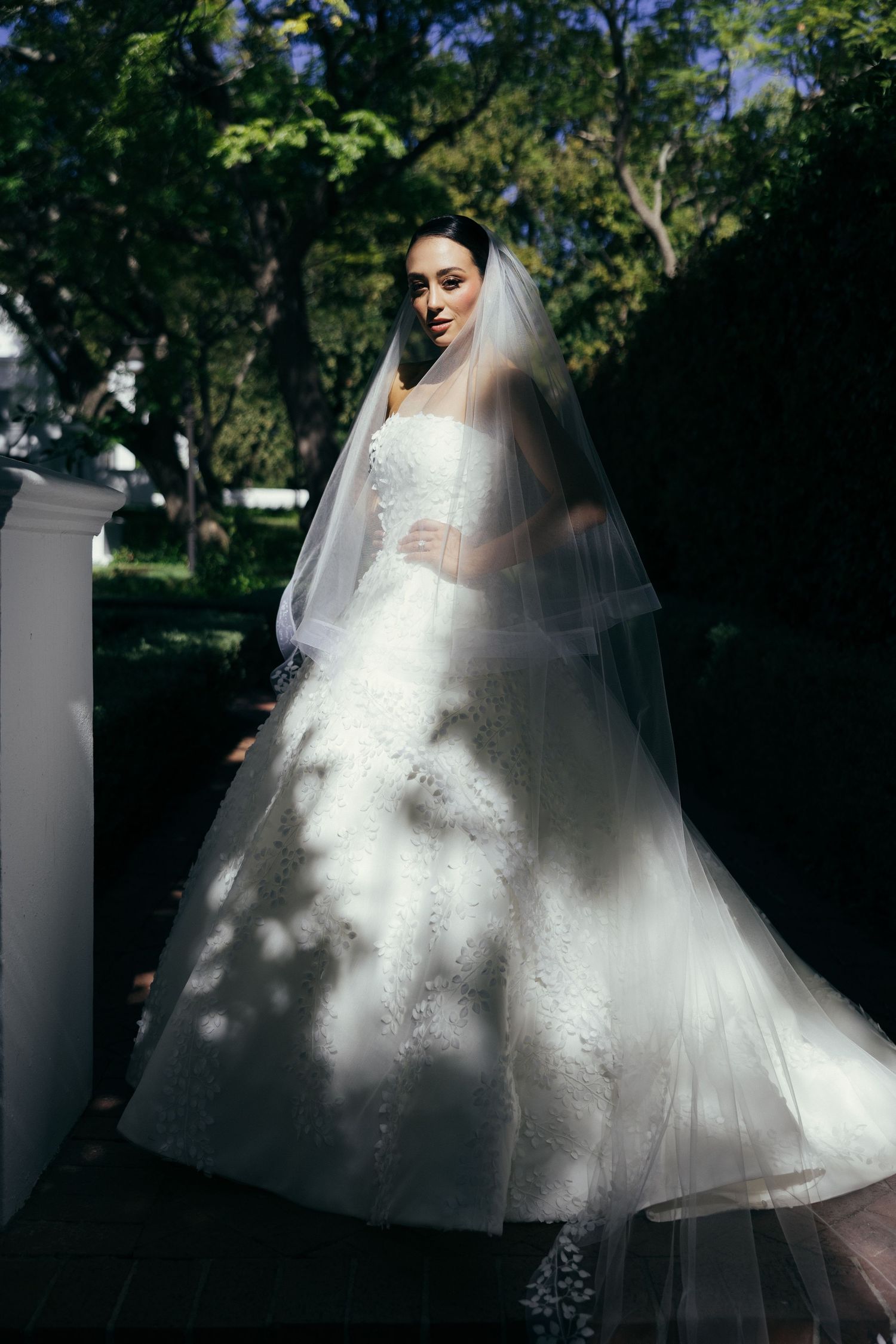 Dramatic bridal portrait featuring full ballgown and veil in dappled garden light.