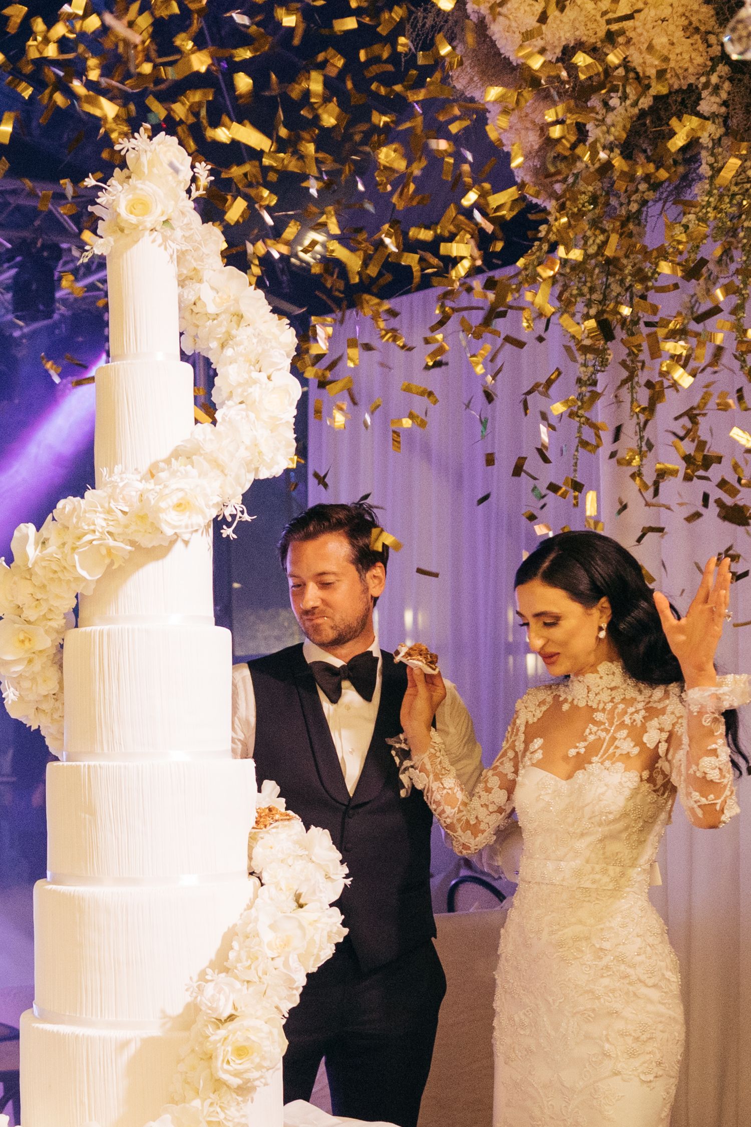 Elegant white tiered wedding cake with cascading flowers and golden confetti falling.