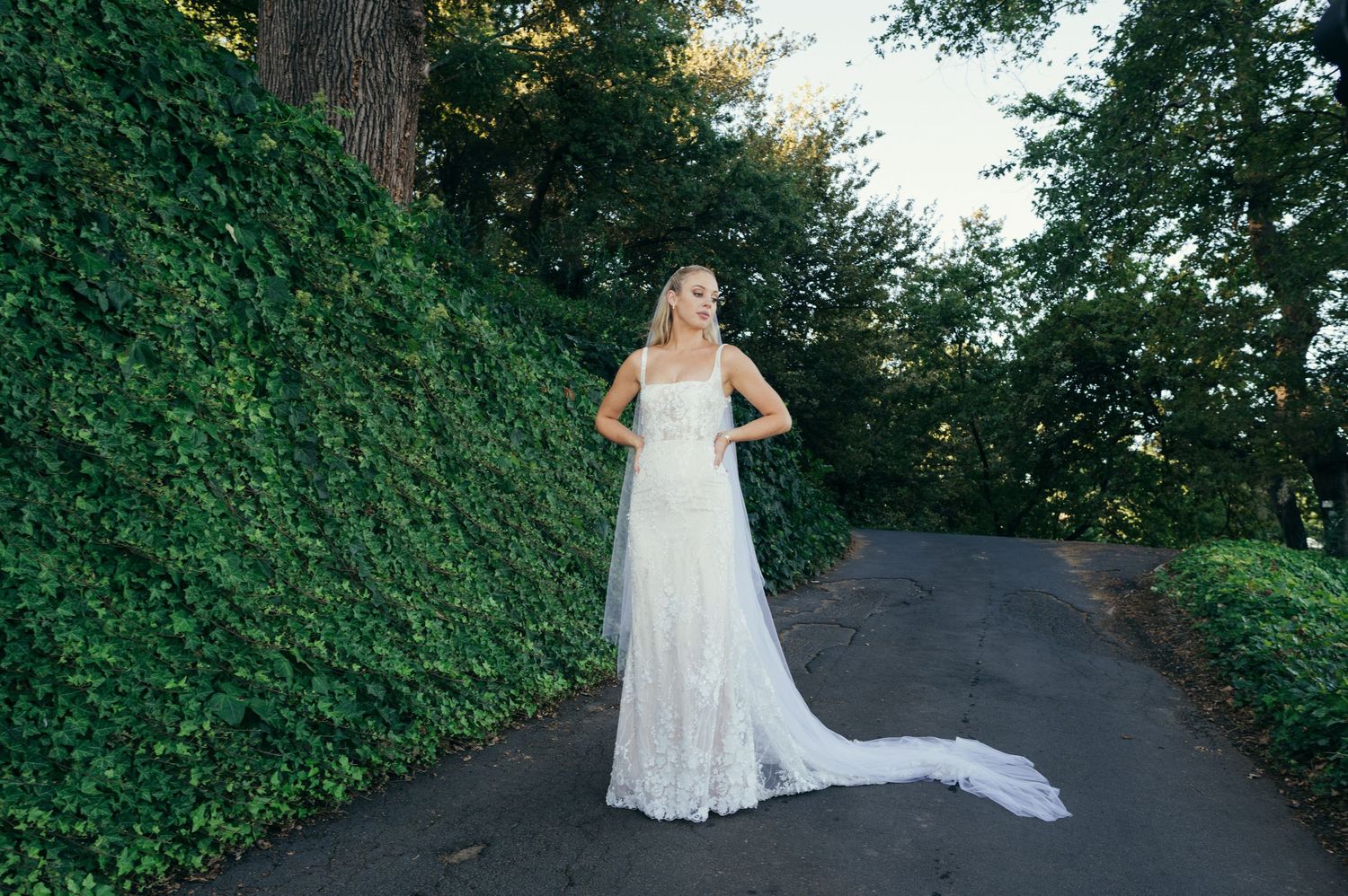 A flowing white wedding gown trails behind on a tree-lined asphalt path.
