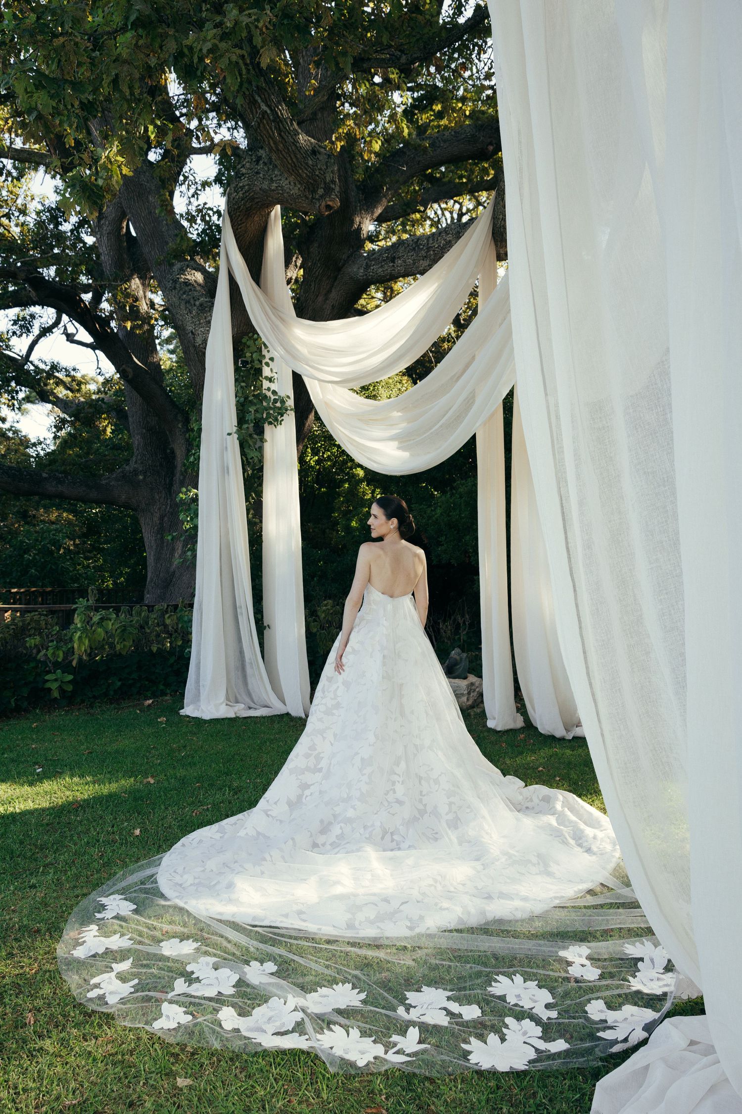 Stunning bride in cathedral length lace train poses under elegant white fabric draping in garden setting.