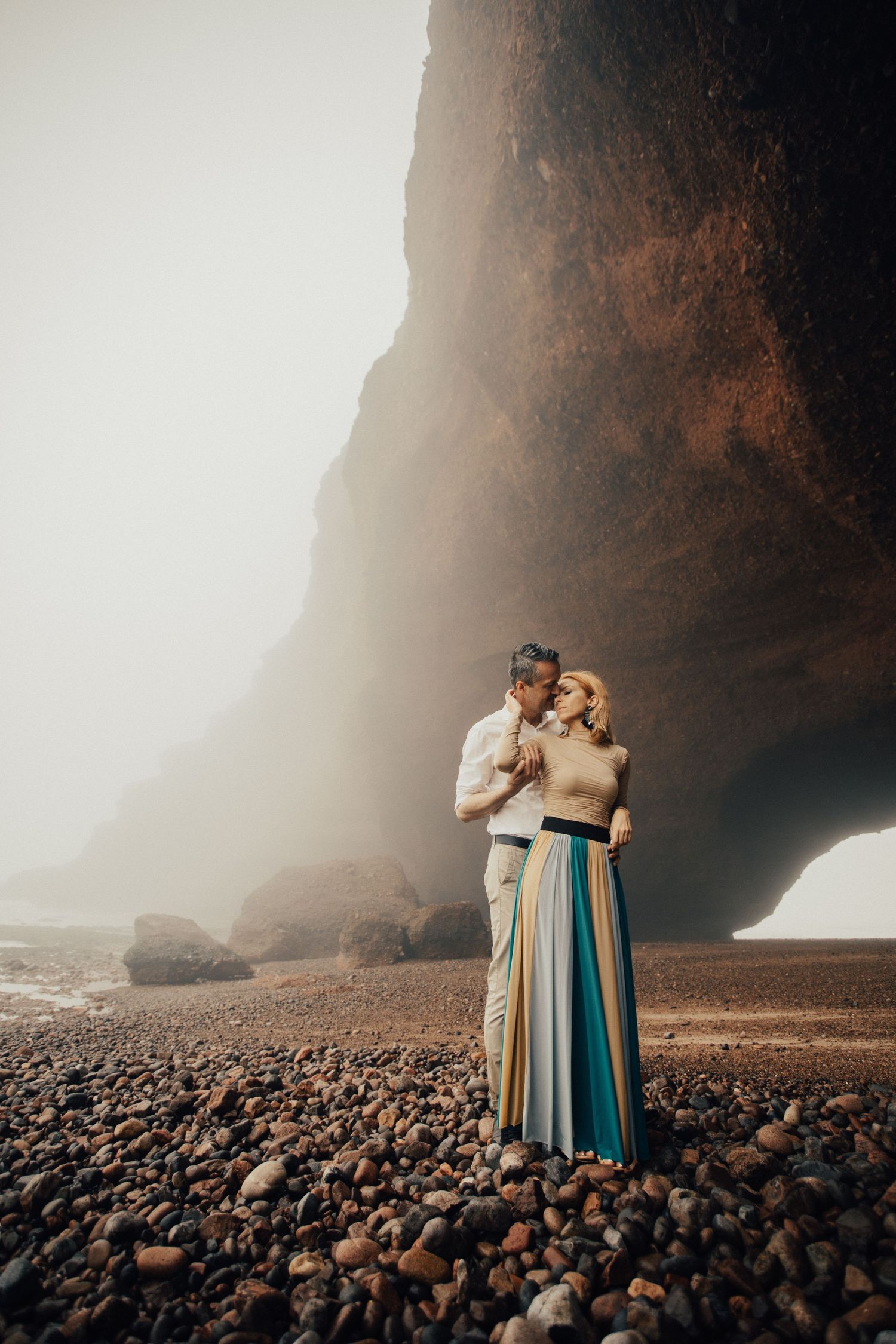 Romantic couple embraces on rocky beach with dramatic sea cliffs and moody lighting in Iceland.