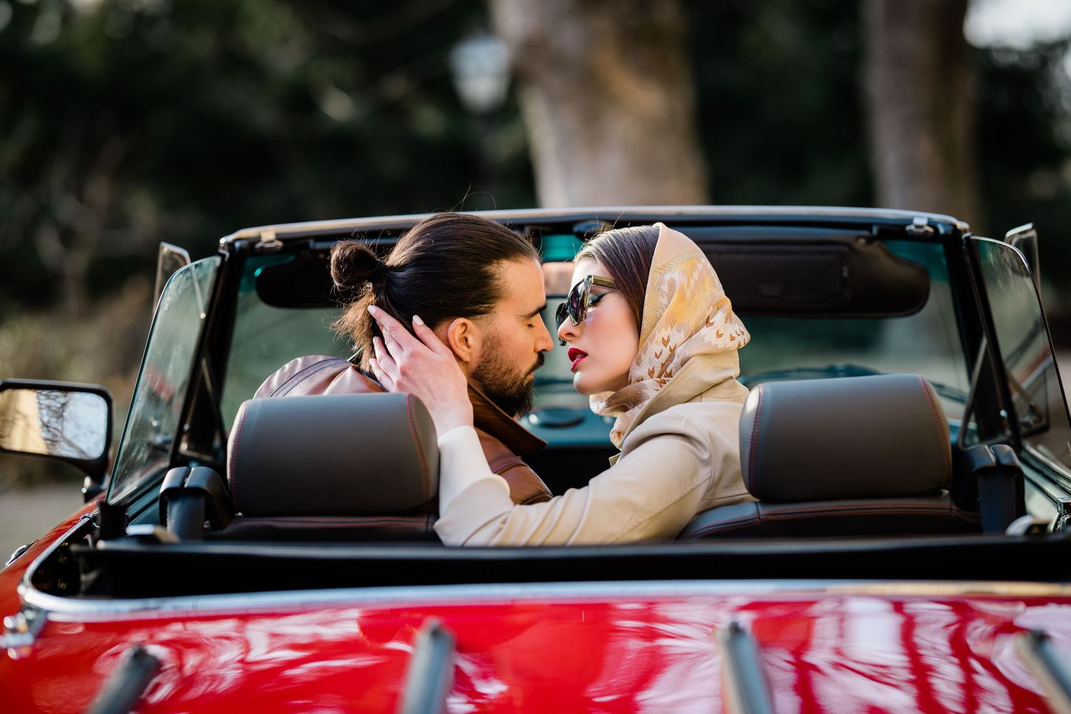 A couple shares an intimate moment together while sitting in a red vintage convertible car on a sunny day.
