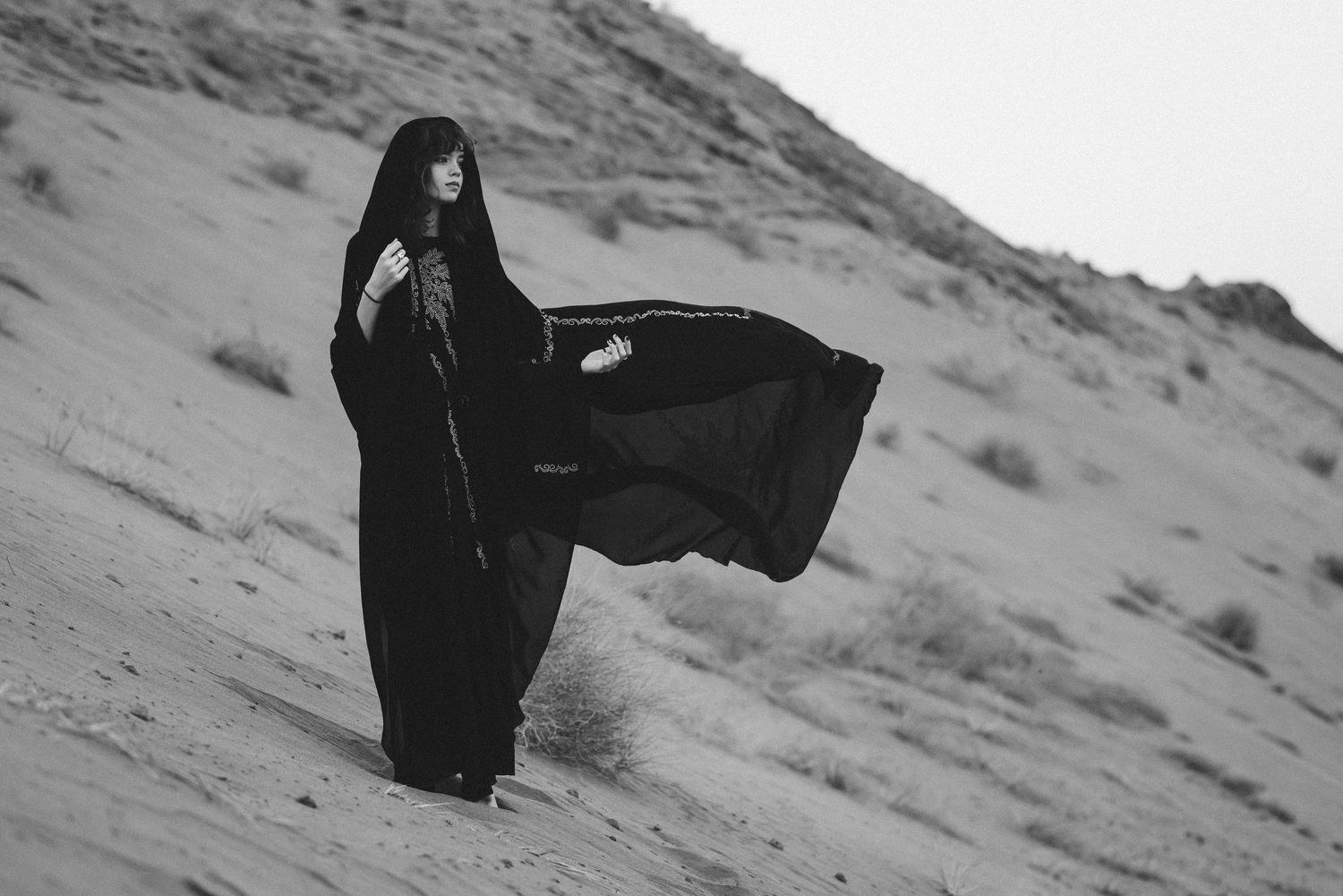A person in dark clothing stands on a desert hillside against an overcast sky.