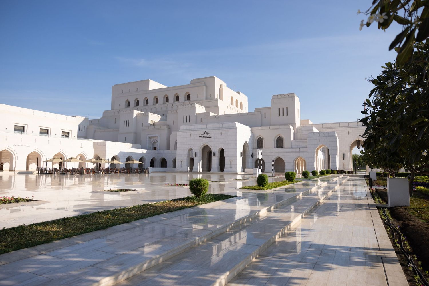 A Muscat-i Royal Opera House fehér márvány architektúrával és tükröződő medencékkel, ragyogó kék ég alatt, Ománban.