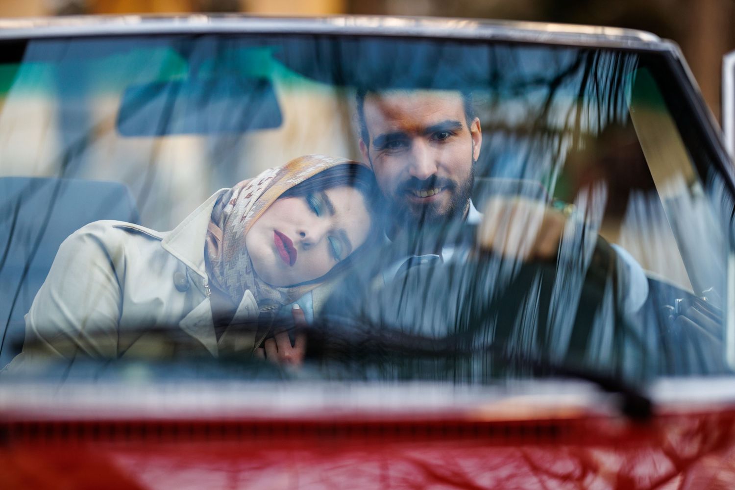A couple sits together in the front seat of a red car viewed through a rain-streaked windshield.