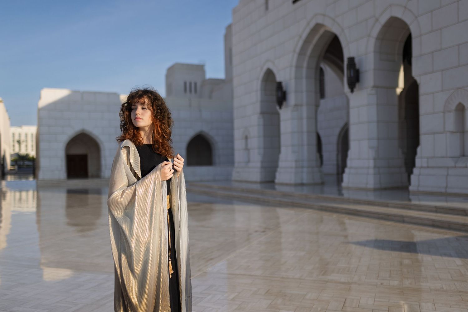 A person in a white dress stands in front of ornate white marble arches at a grand mosque or palace.