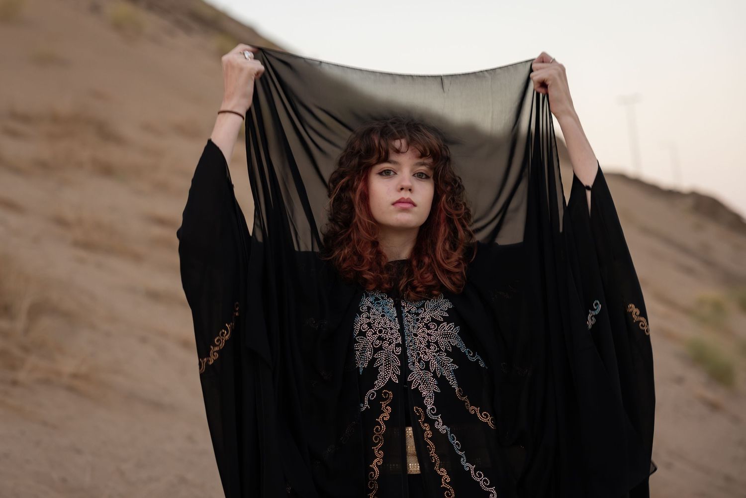 A figure in a black flowing dress stands against a rustic wall in dramatic lighting.