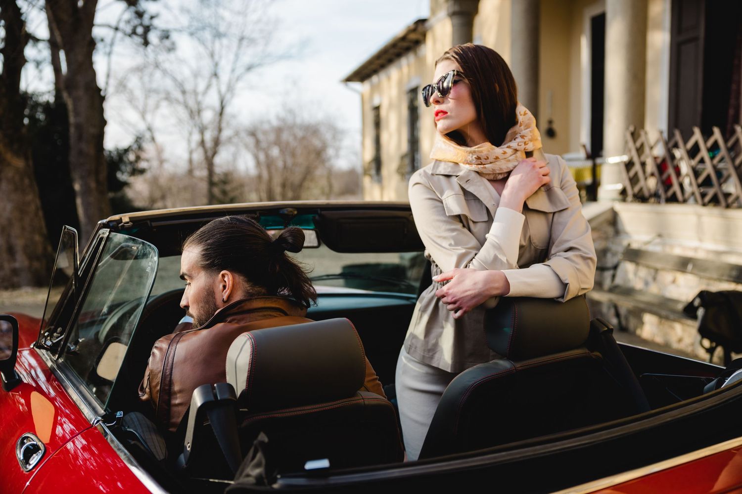 Two people sitting together in a black vintage convertible car parked outside a classical building.