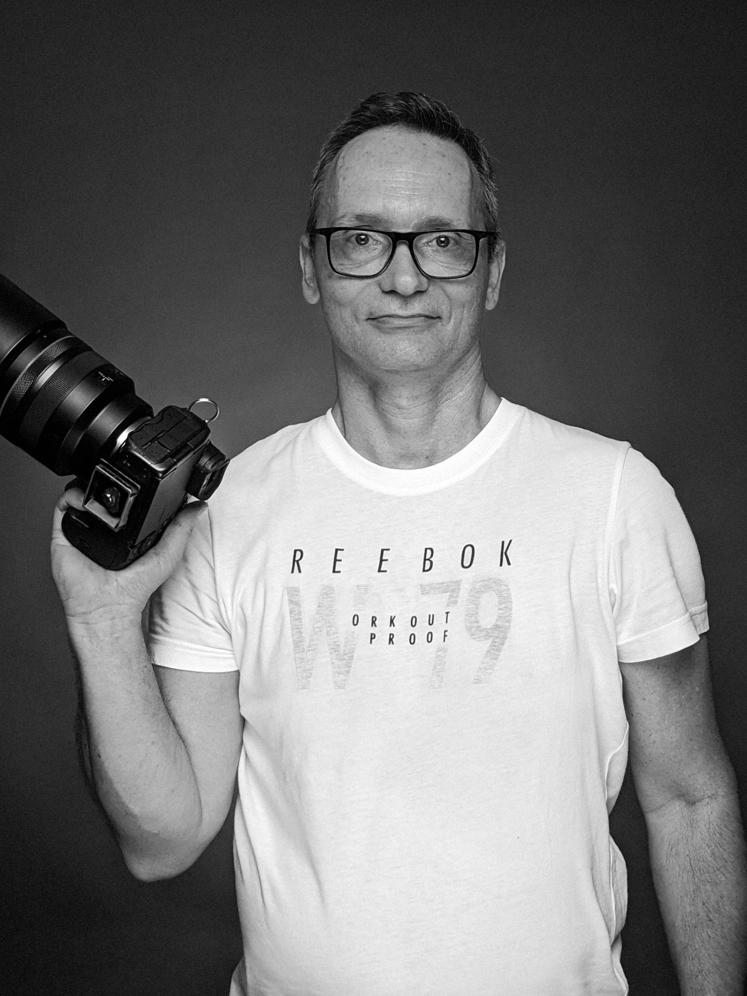 Professional photographer wearing glasses and a white Reebok t-shirt holds a camera against dark background.