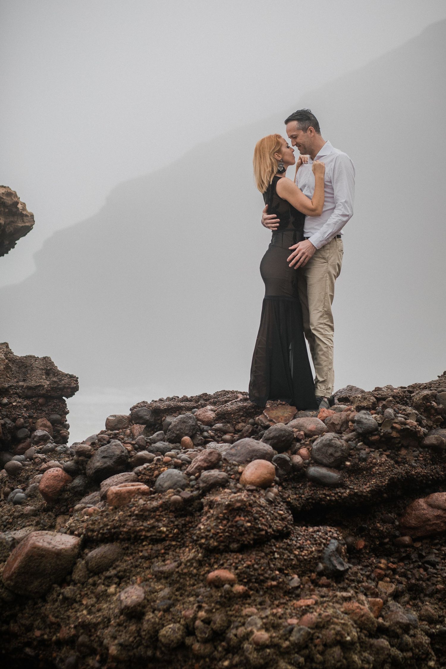 A couple embraces atop a rocky volcanic terrain on a misty overcast day.