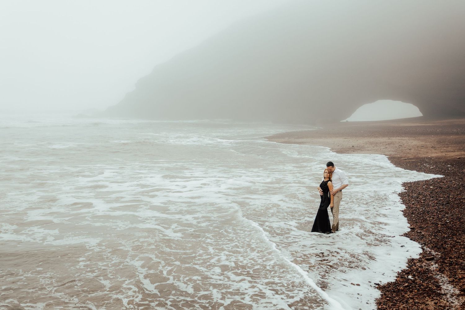 Silhouetted figure stands on a misty rocky beach at sunset with waves crashing against the shore.