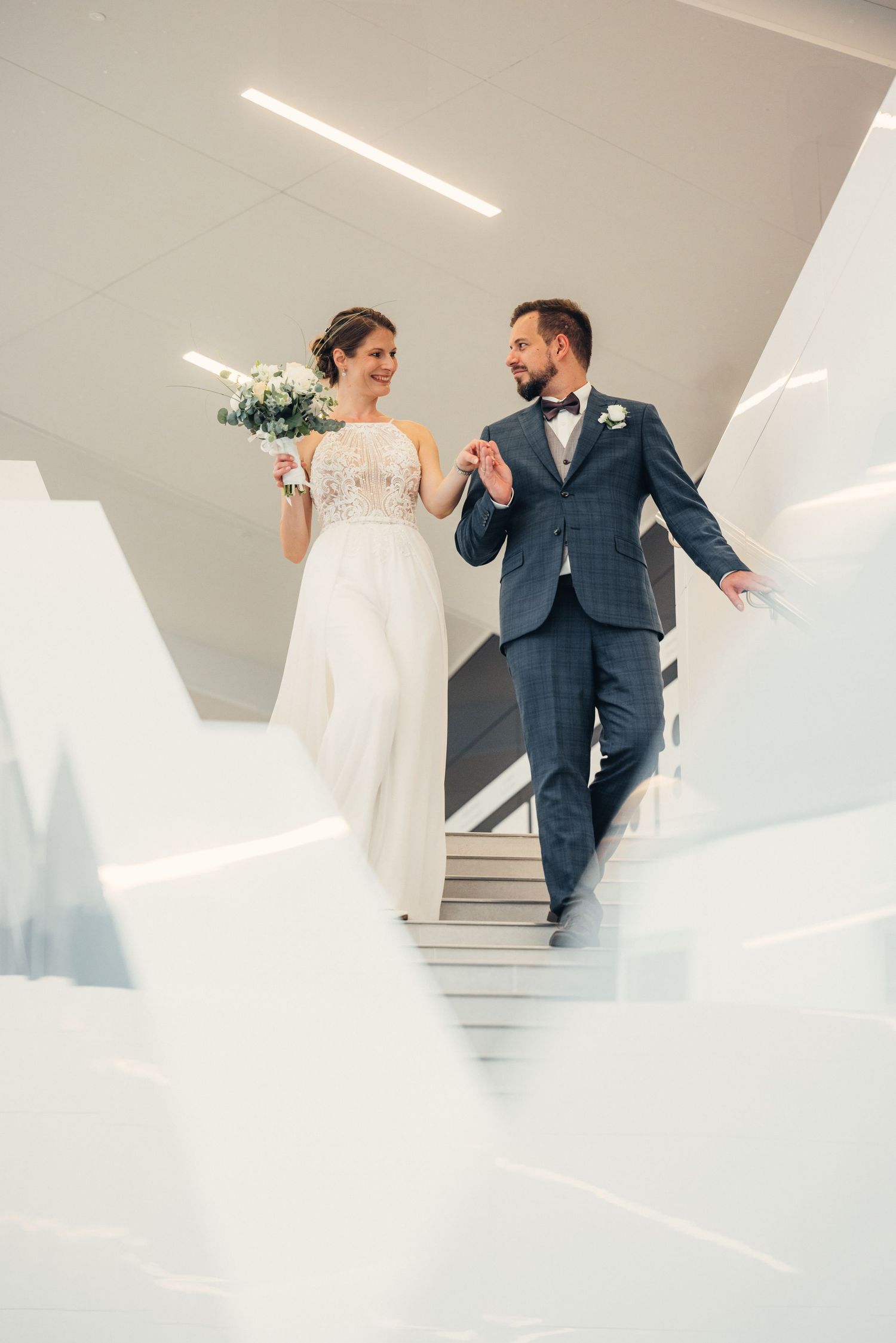 Elegáns pár ereszkedik le a fehér lépcsőn esküvőjük napján modern építészeti környezetben.