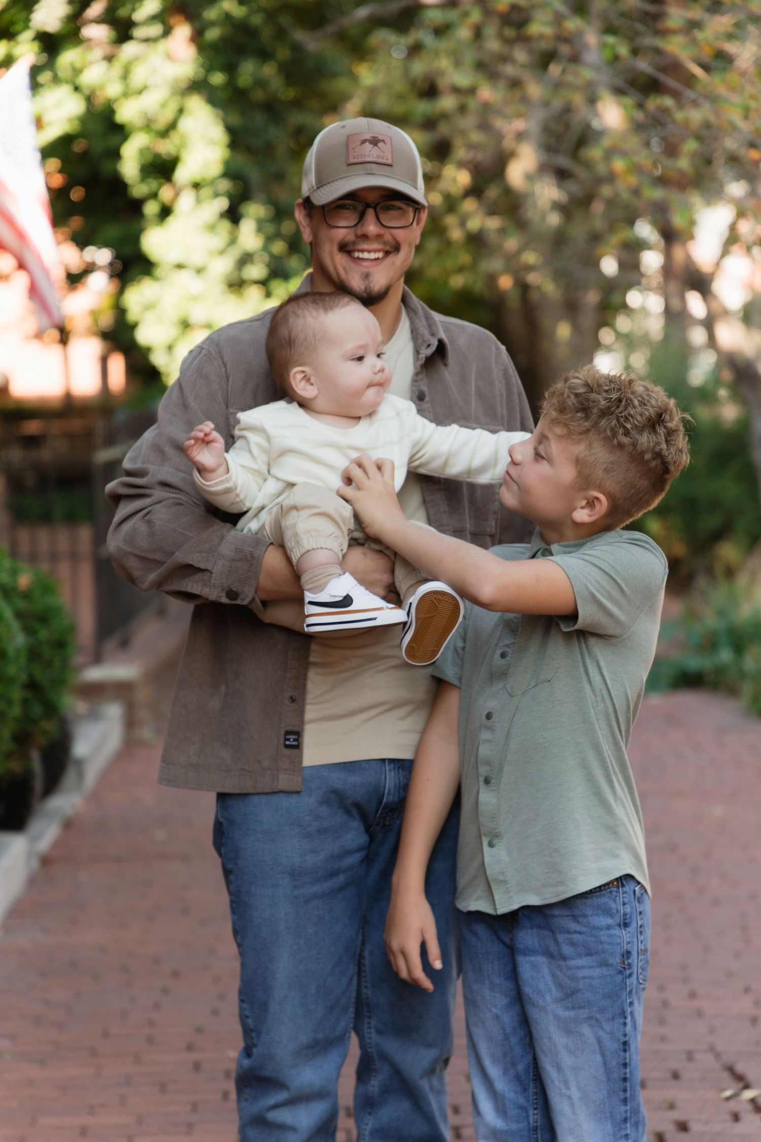 A smiling man in a cap holds a baby while an older boy reaches up to interact with the infant outdoors.
