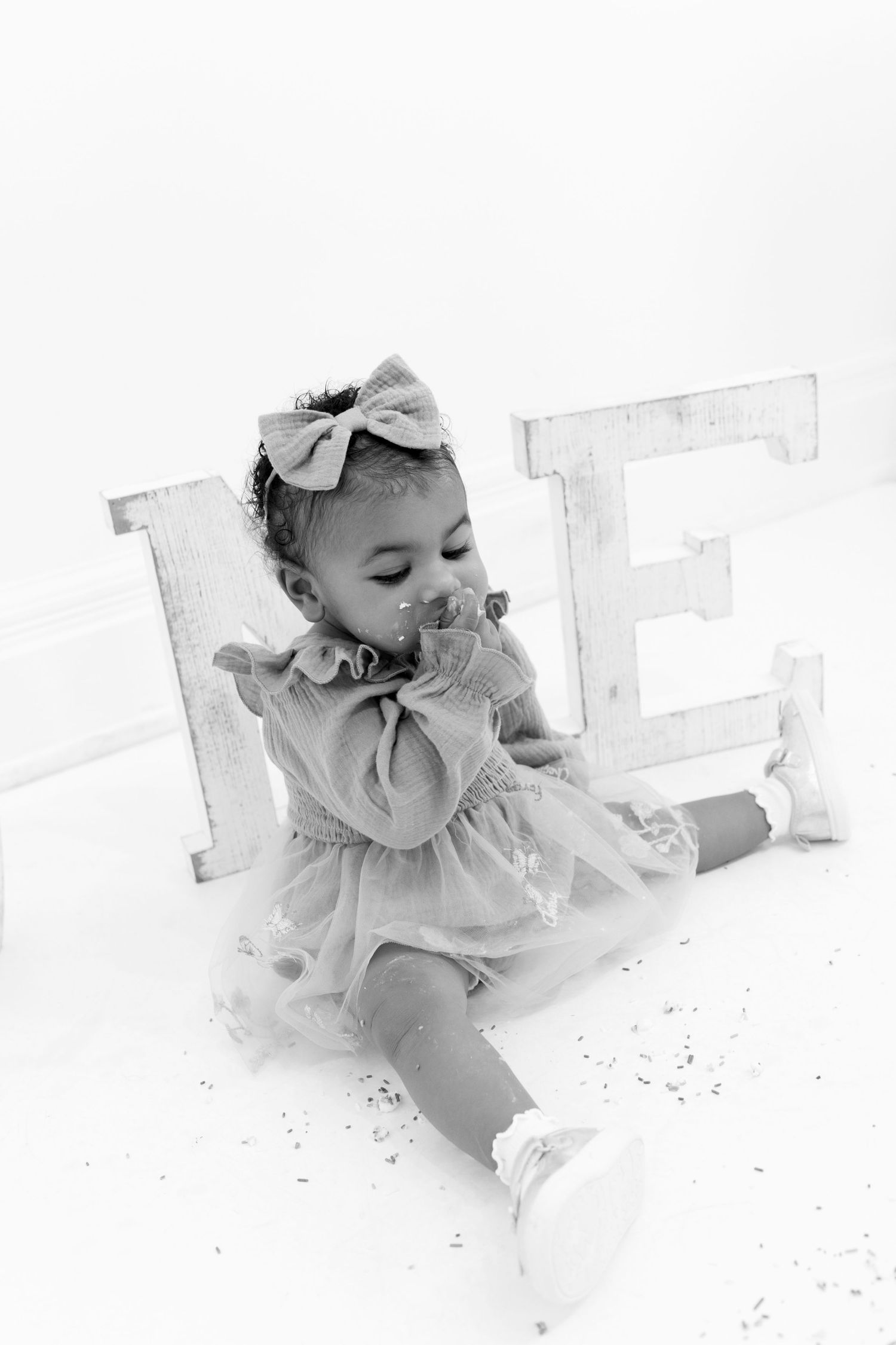 Black and white photo of a baby girl in a tutu dress with a bow headband sitting by decorative letters.