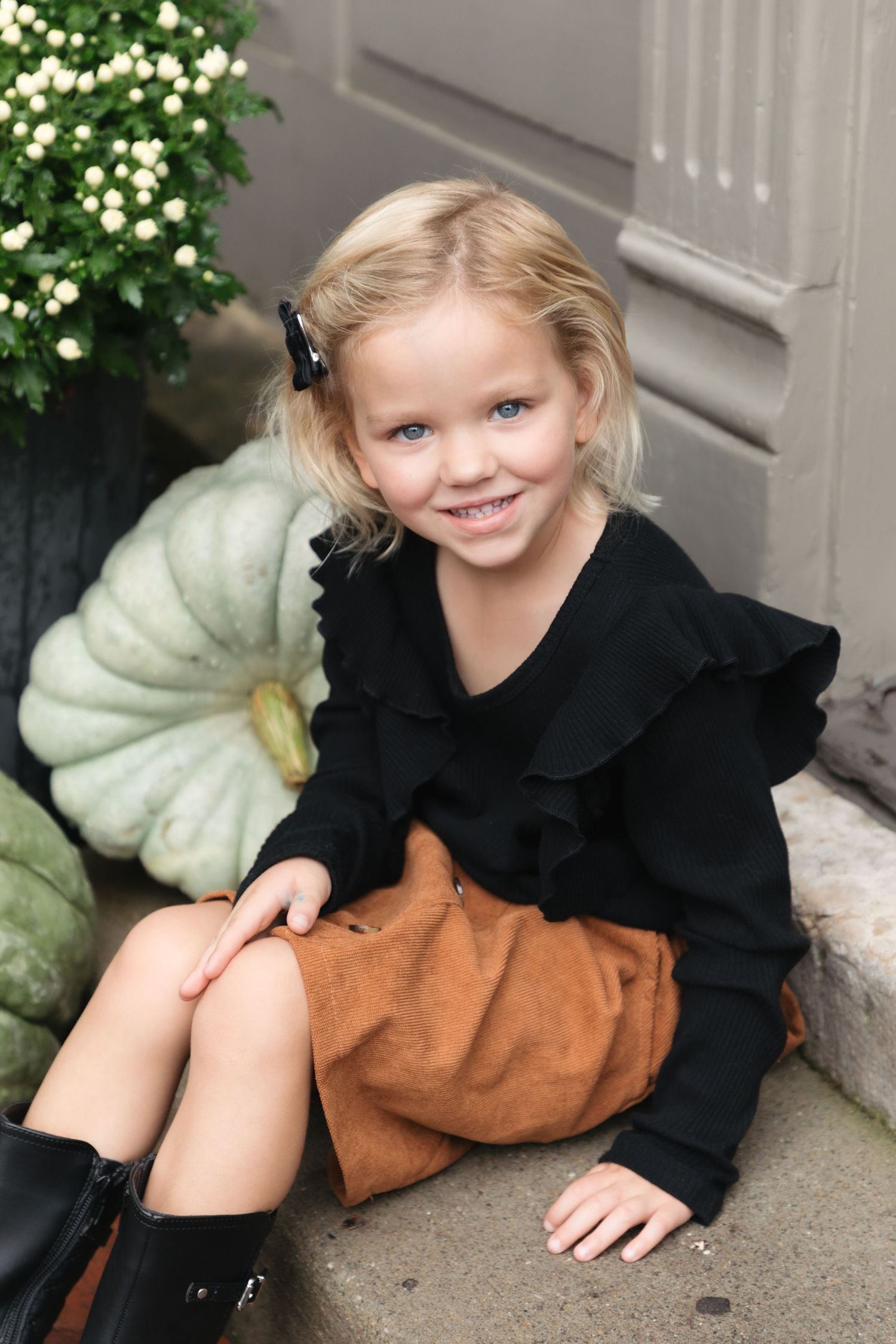Smiling blonde girl in black ruffled top, brown skirt, and boots sits on steps beside green pumpkins in fall setting.