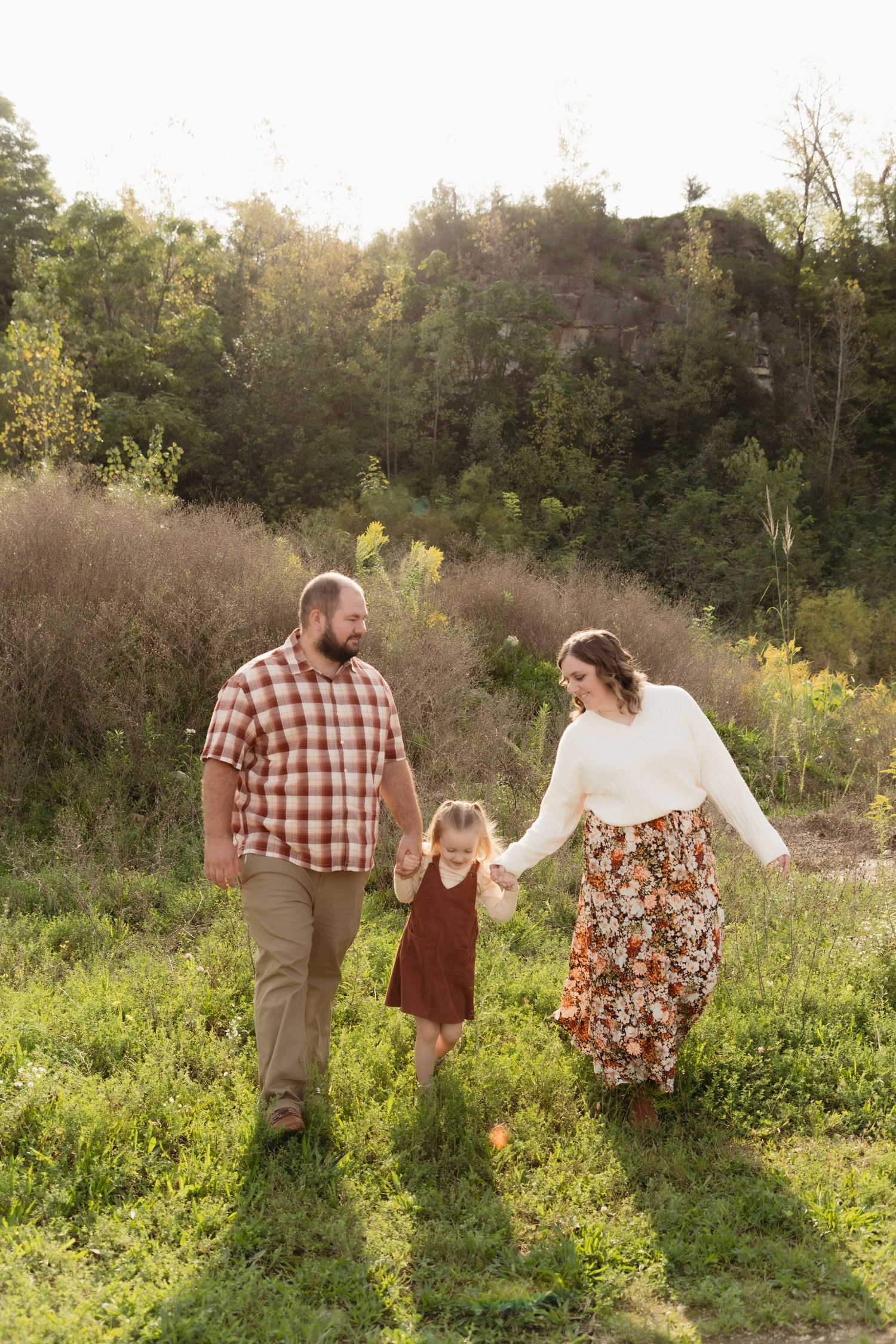 A family of three walks hand-in-hand through a sunlit meadow, parents guiding their young daughter in autumn outdoor setting.