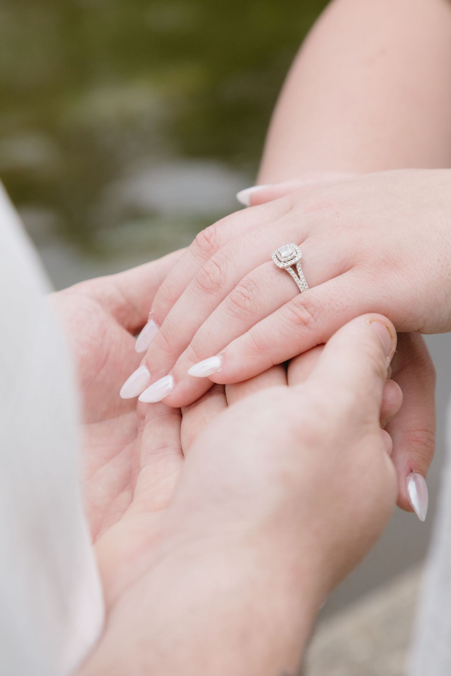 Close-up of hands holding, featuring a diamond halo engagement ring on a finger with white almond-shaped nails.