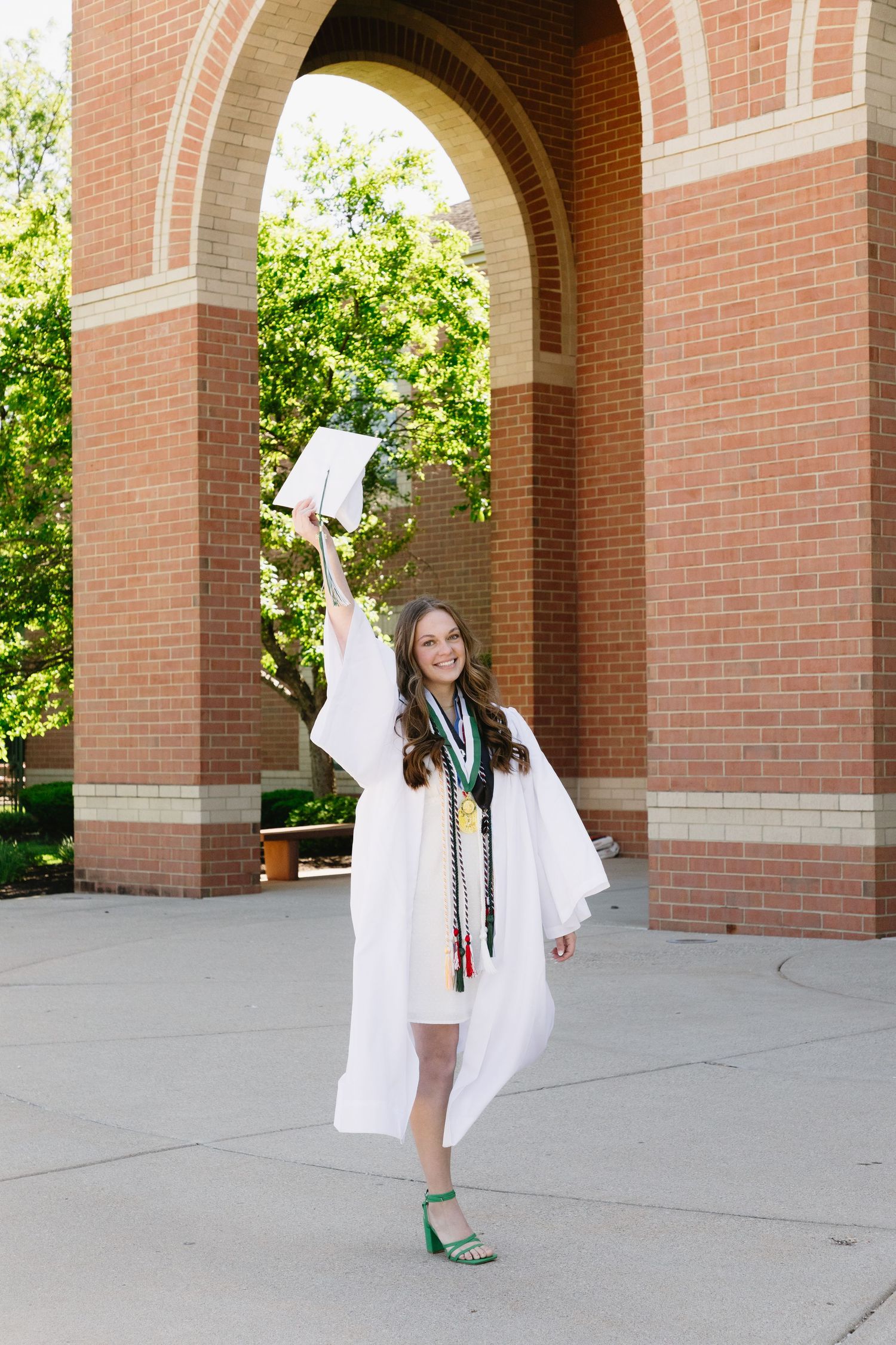 A smiling graduate in white cap and gown raises her diploma outside a brick arch, celebrating her graduation day.