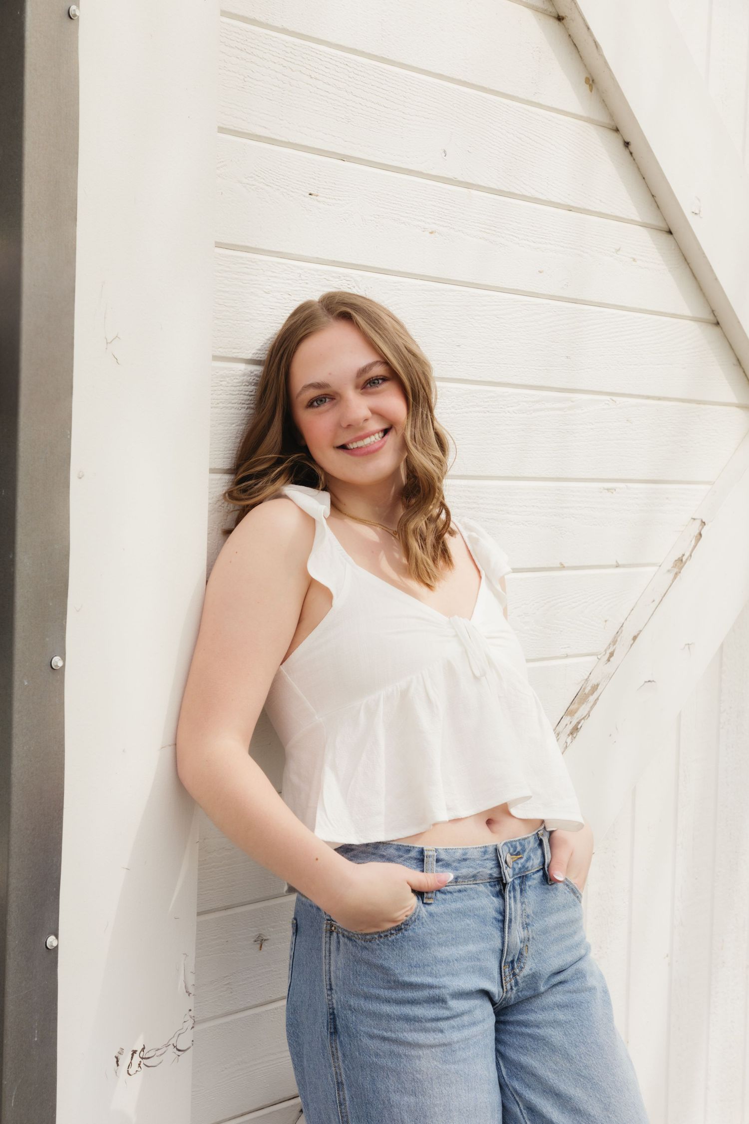 Smiling young woman in white crop top and jeans leaning against a white wooden wall outdoors.
