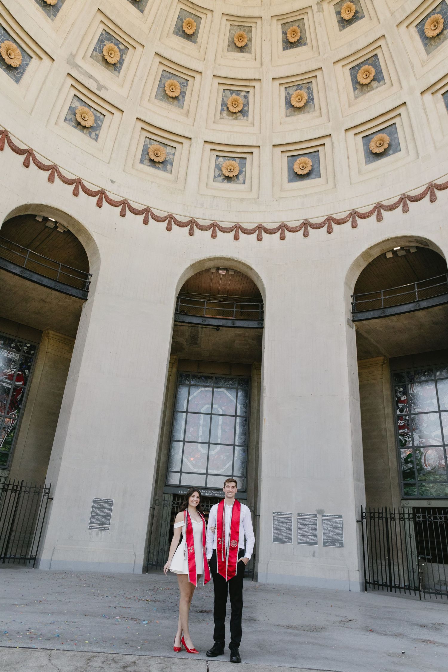 Two graduates in red sashes pose under ornate domed ceiling with arched windows at a neoclassical building.