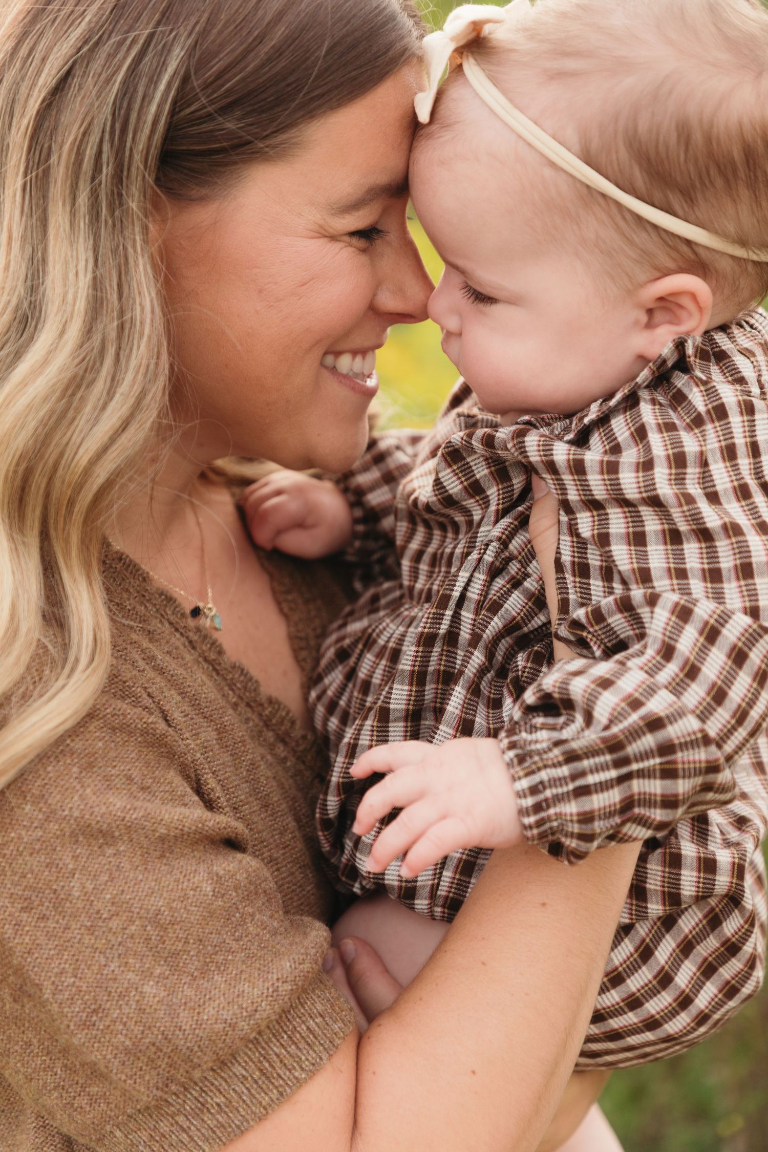 A smiling mother tenderly touches foreheads with her happy baby girl in a plaid dress outdoors.