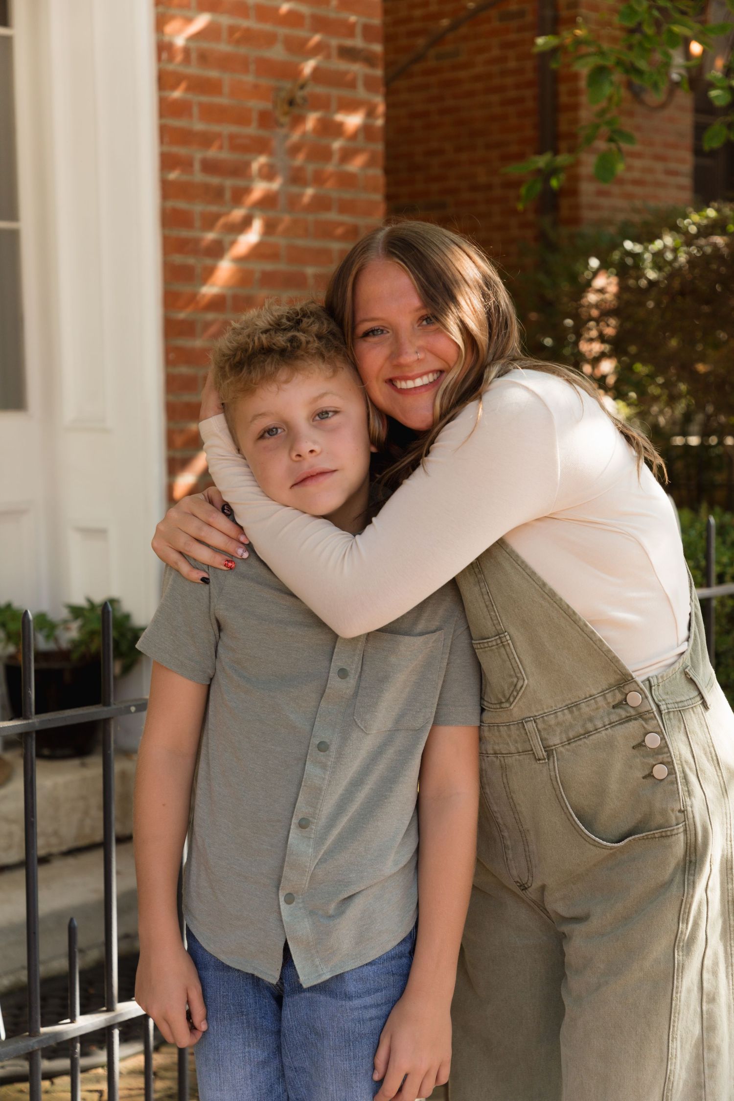 A smiling teenage girl hugs a young boy in front of a brick building outdoors.