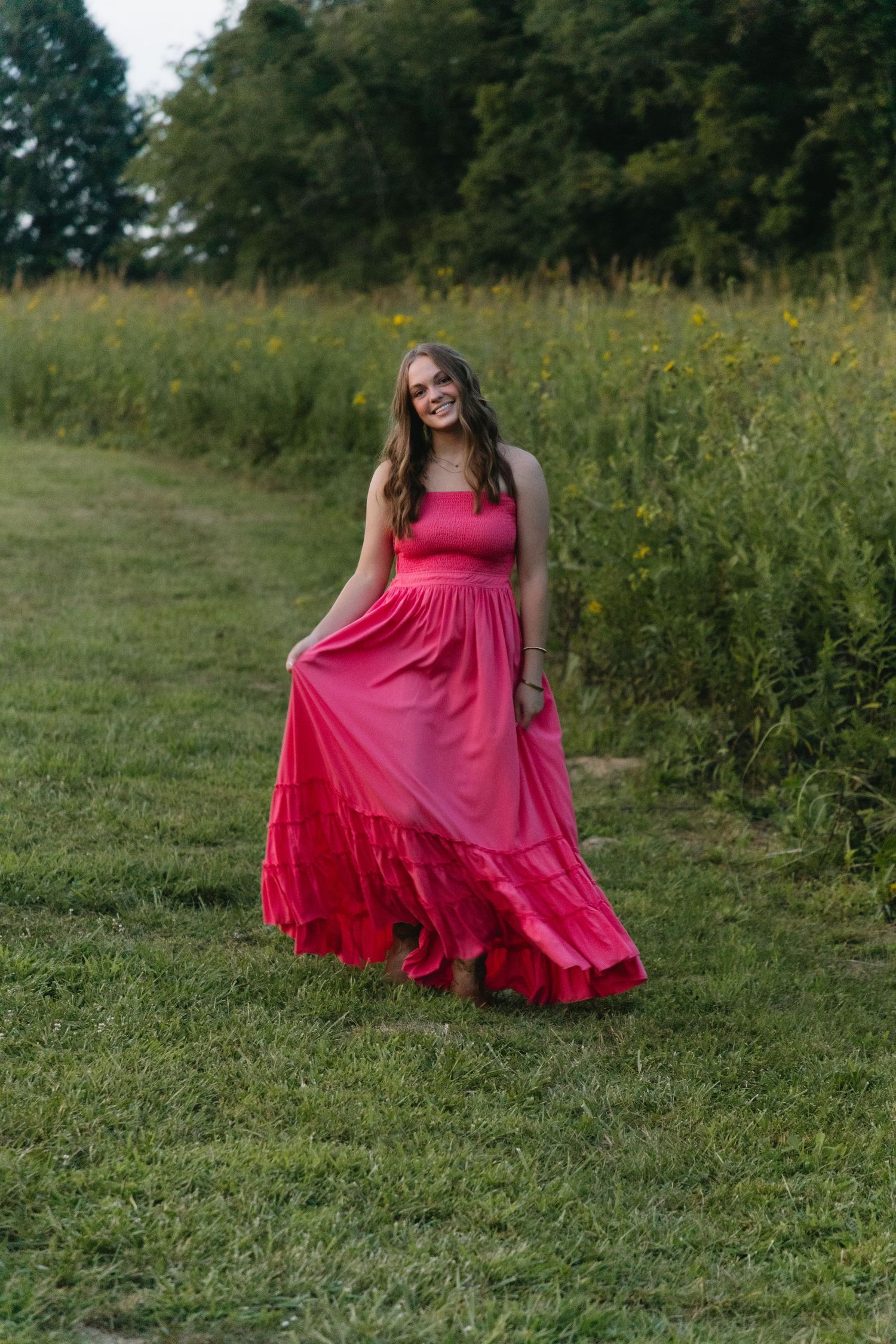 A woman in a flowing red maxi dress smiles while standing in a lush green field with wildflowers.