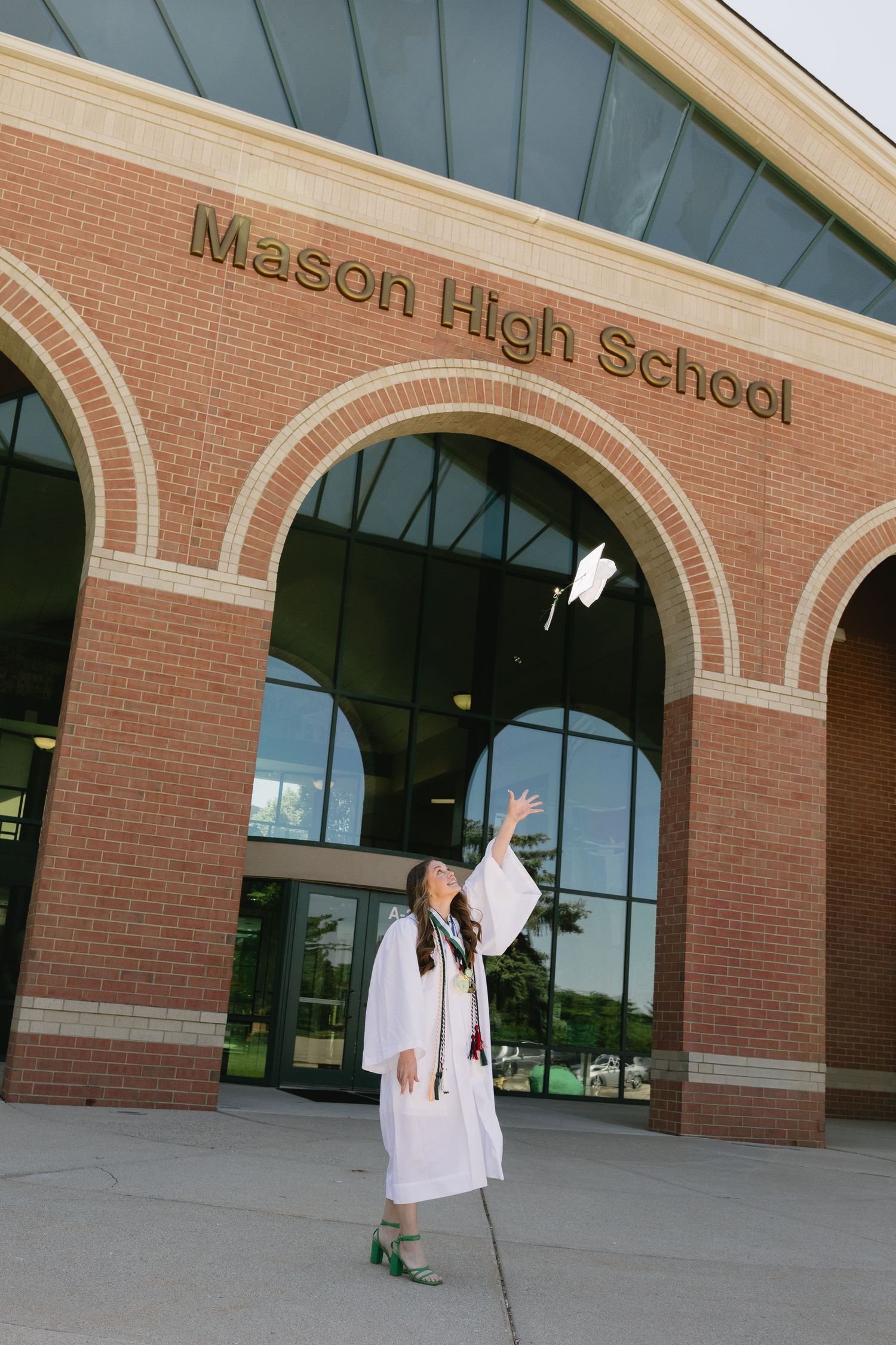 A graduate in white cap and gown tosses her cap in front of Mason High School brick building.